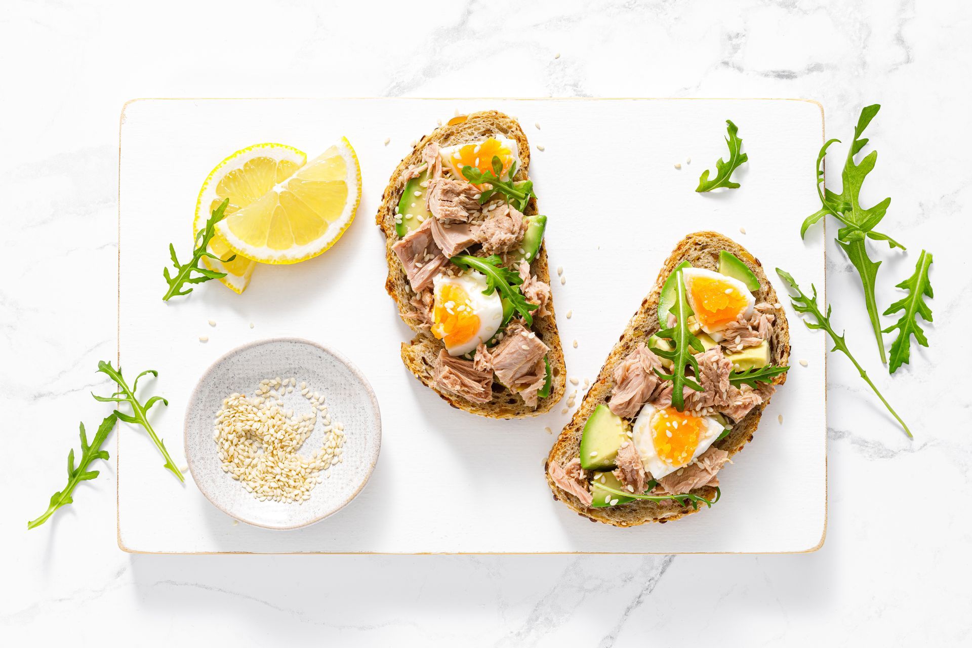 Two open-faced sandwiches topped with tuna, avocado, and egg, garnished with arugula and lemon, on a white cutting board.