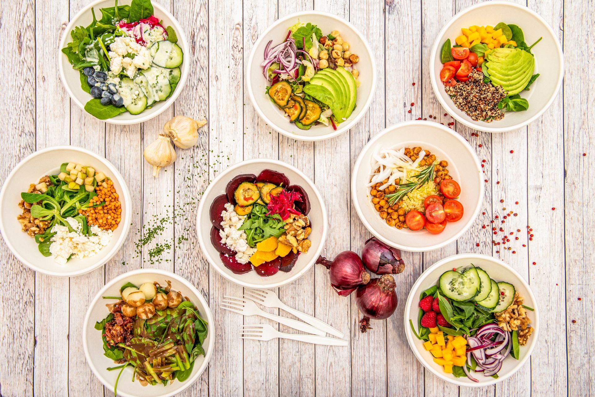 Various colorful salad bowls on a white wooden table, including vegetables, grains, and fruits.