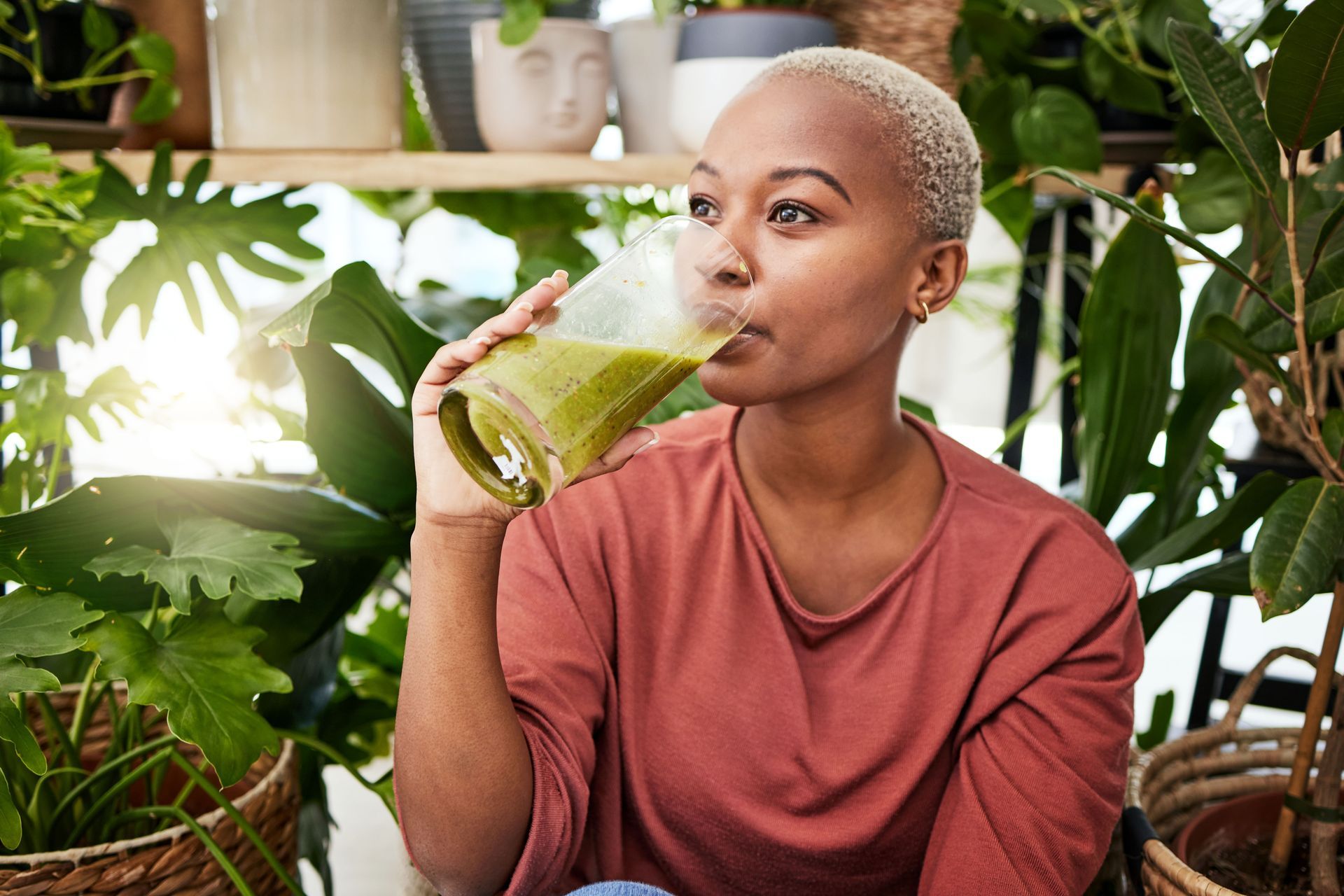 Woman with short blonde hair sipping a green smoothie surrounded by lush indoor plants. She wears a red shirt and appears relaxed.