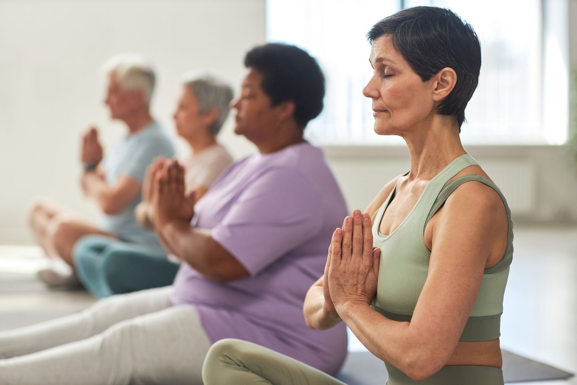Group of diverse seniors in yoga class, sitting in lotus position with hands in prayer pose, eyes closed. Indoors, with natural light.