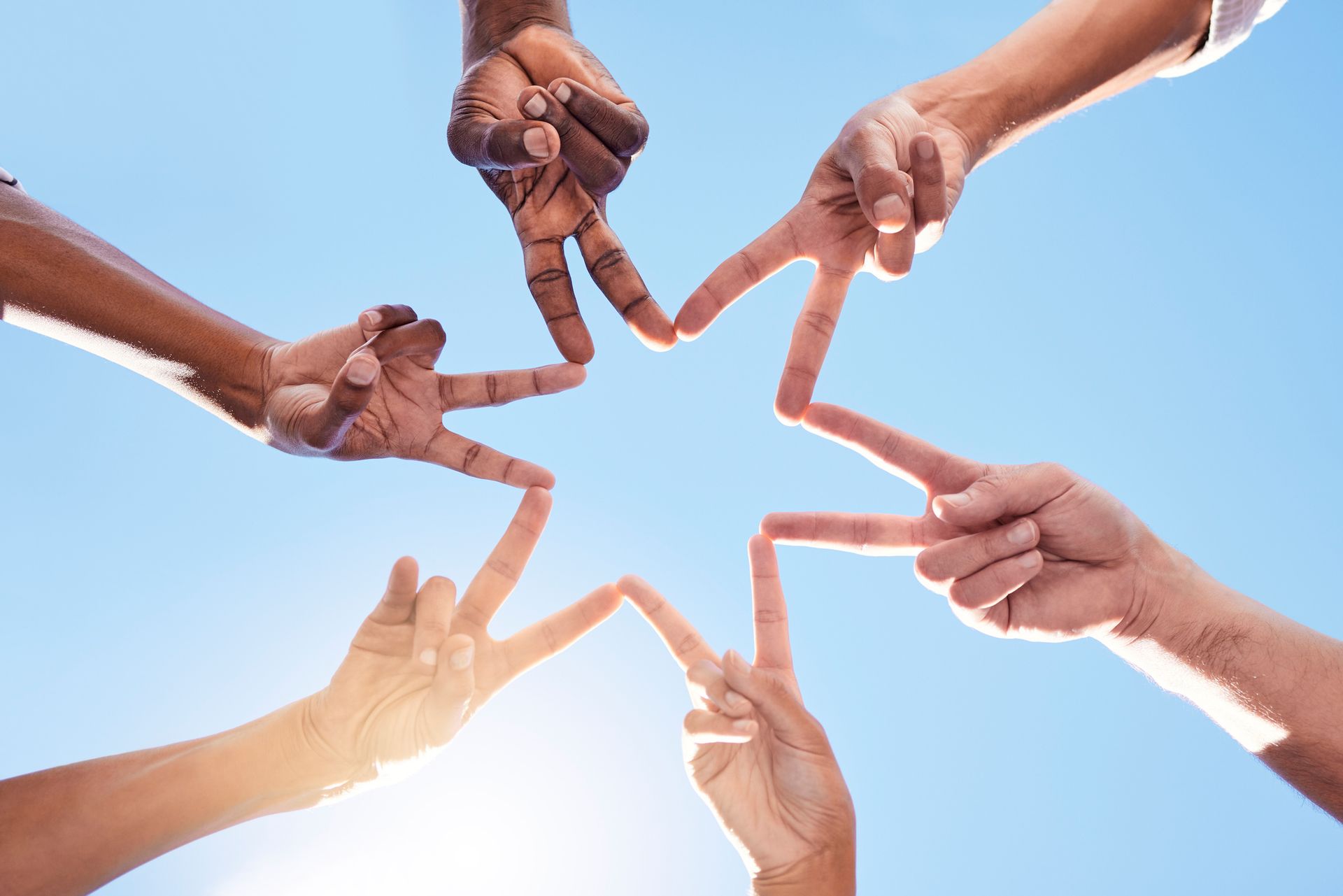 Hands of various skin tones forming a star shape against a blue sky, emphasizing unity and diversity.