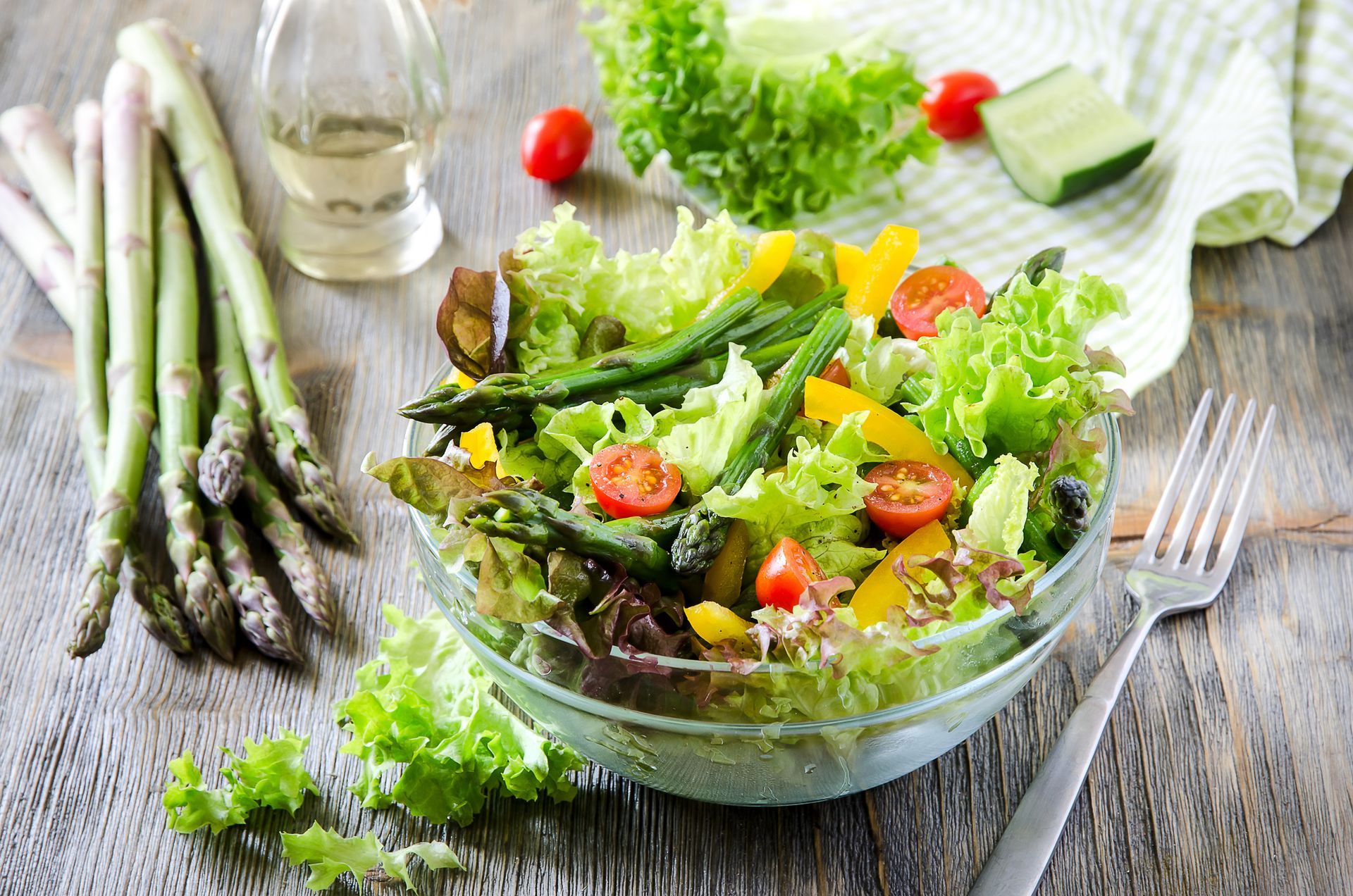 A glass bowl overflowing with a fresh salad, asparagus stalks to the side, tomatoes, and a fork.