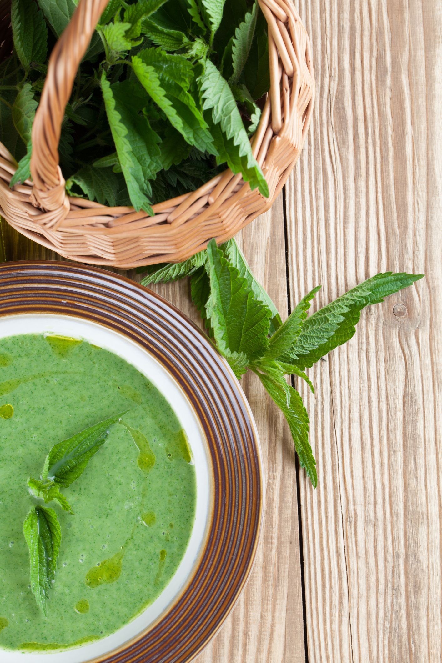 Bowl of green soup with nettle garnish, beside a basket of nettles, set on a wooden surface.