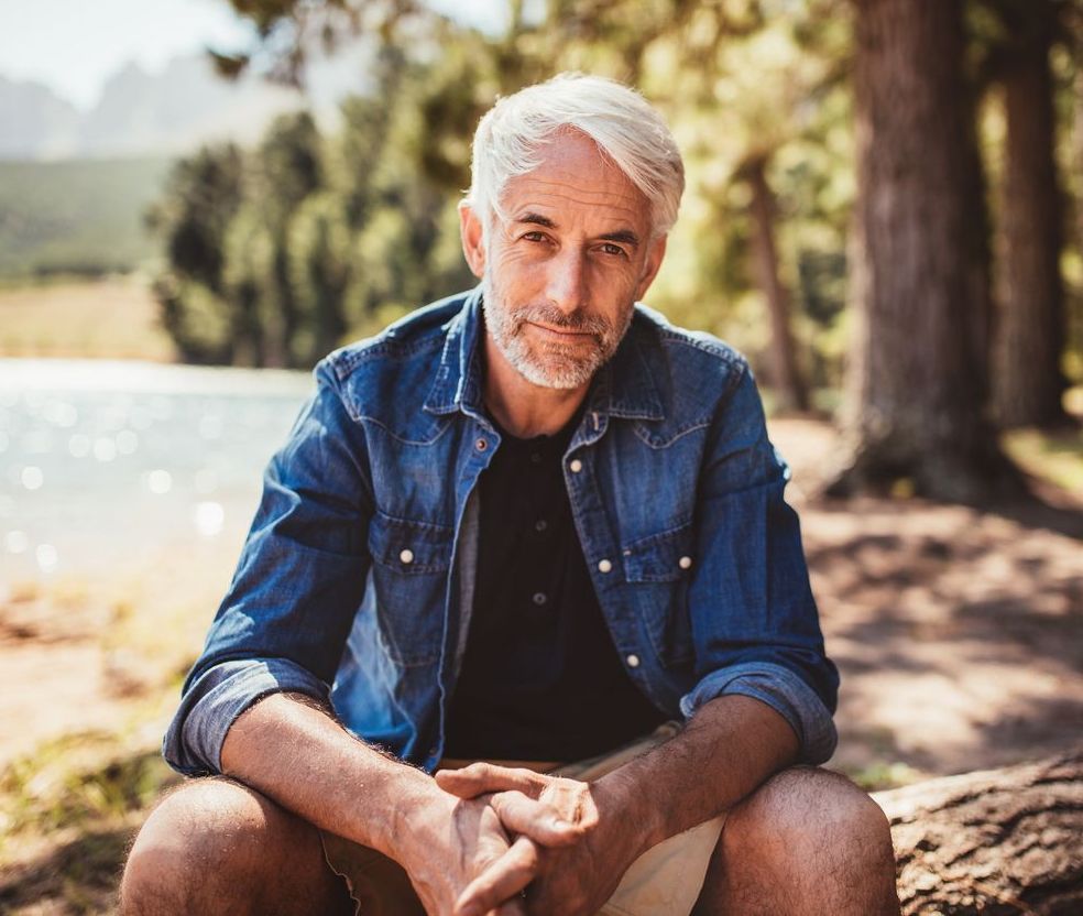 Older man with gray hair, sitting outside near a lake. He's wearing a denim shirt and looking at the camera with a neutral expression.