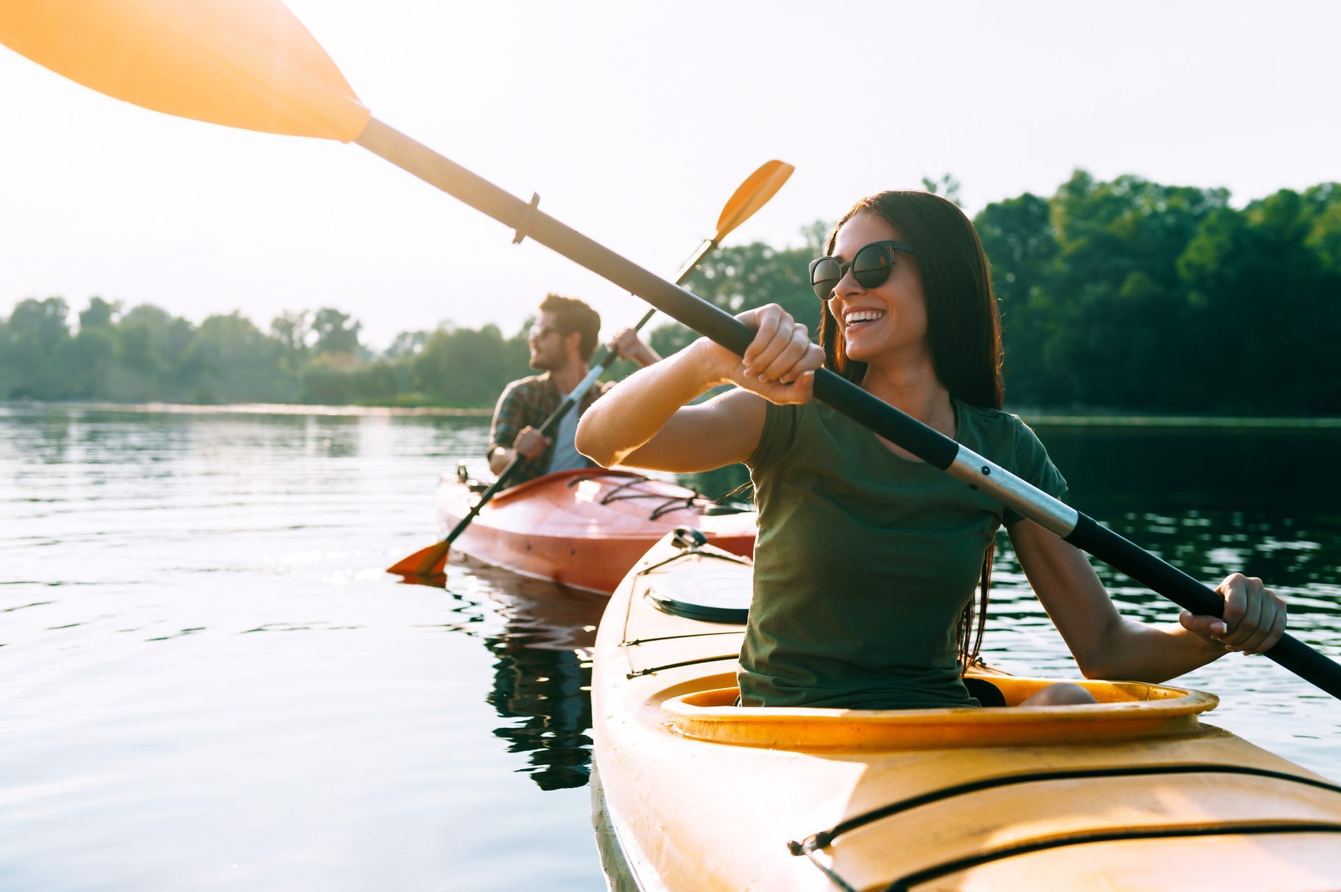 Woman kayaking on a lake, smiling and holding a paddle. Another person kayaks in the background. Sunlight.