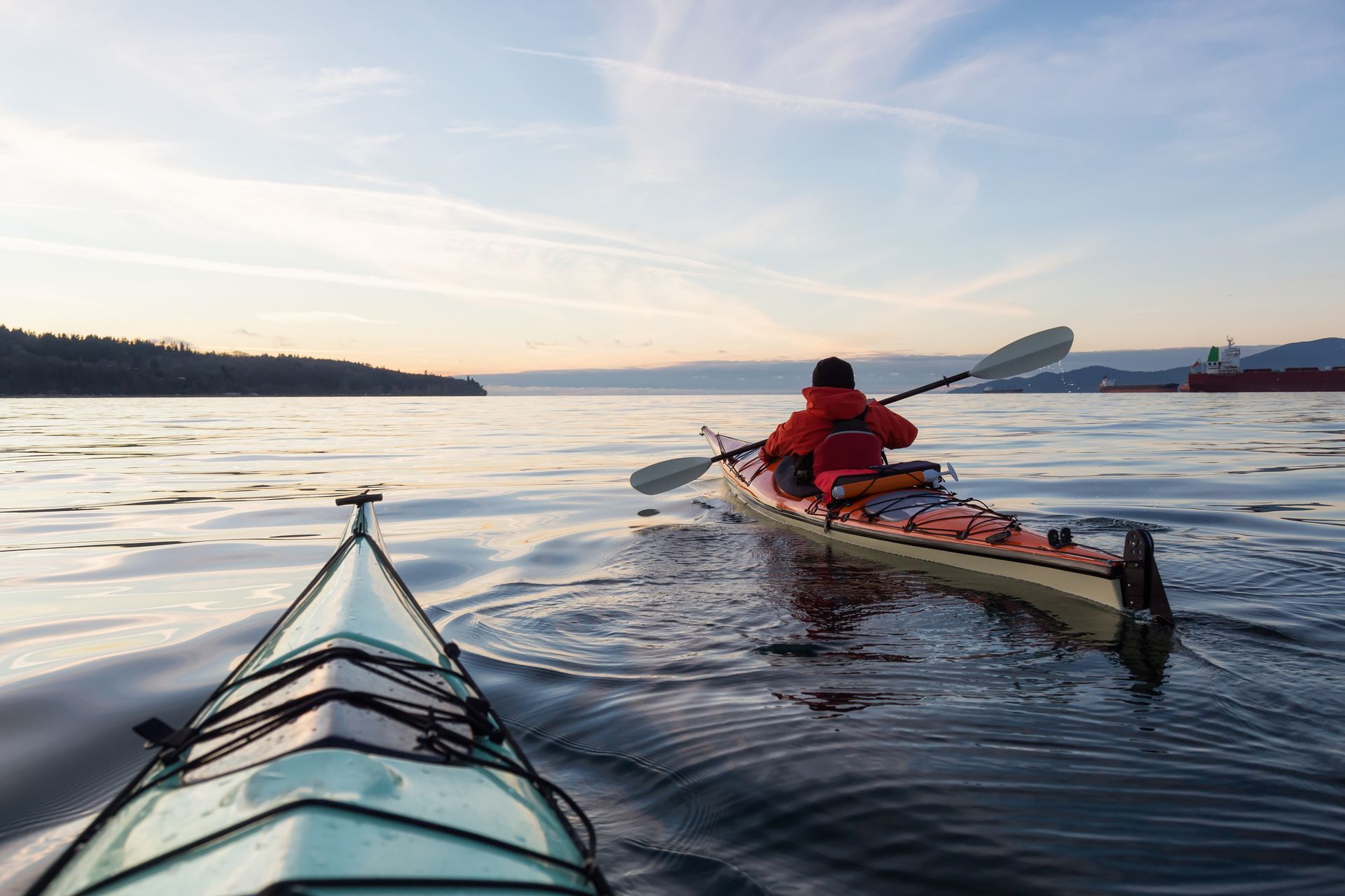 Two kayakers on calm water, one in the foreground, the other paddling away, with a shoreline and a ship in the distance.