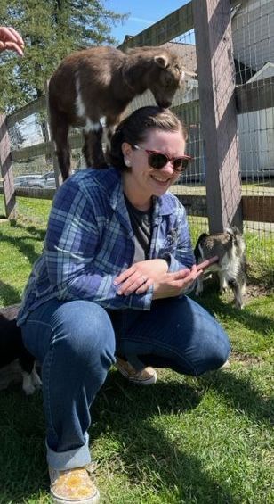 Woman smiles with a goat standing on her head, squatting in a grassy pen. Another small goat approaches her hand.