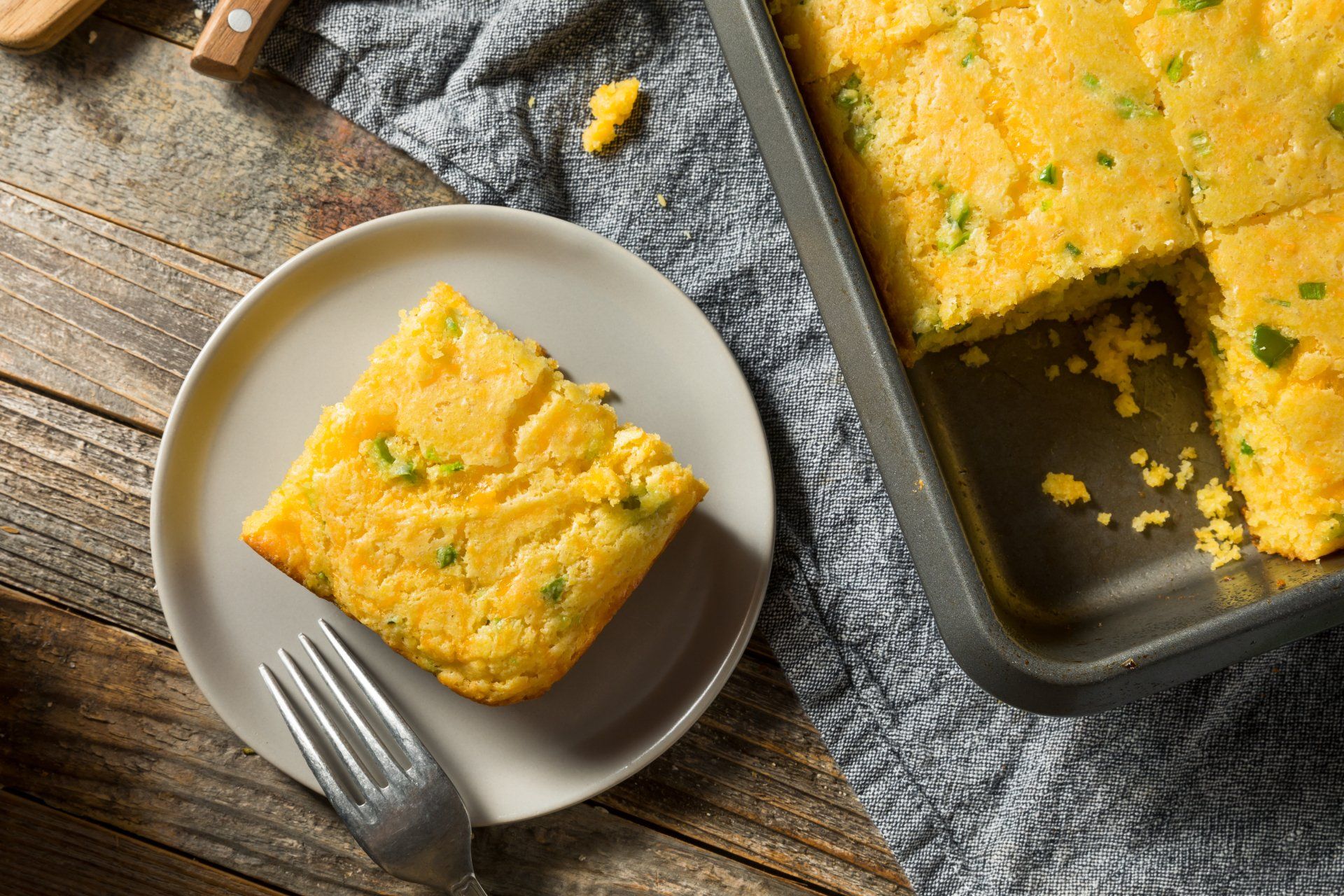Cornbread slice on a plate with a fork, next to a baking pan of cornbread on a wooden table.