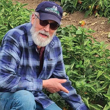 A man in a blue plaid shirt, jeans, and a Tacoma cap points at a green plant in a garden.