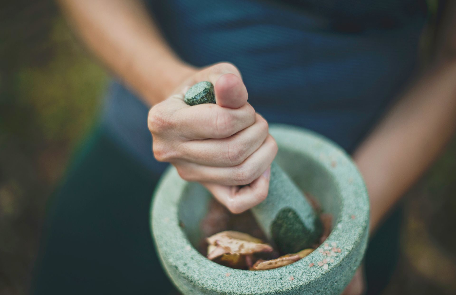 Person grinding herbs in a stone mortar and pestle, outdoors. They are wearing a teal top.