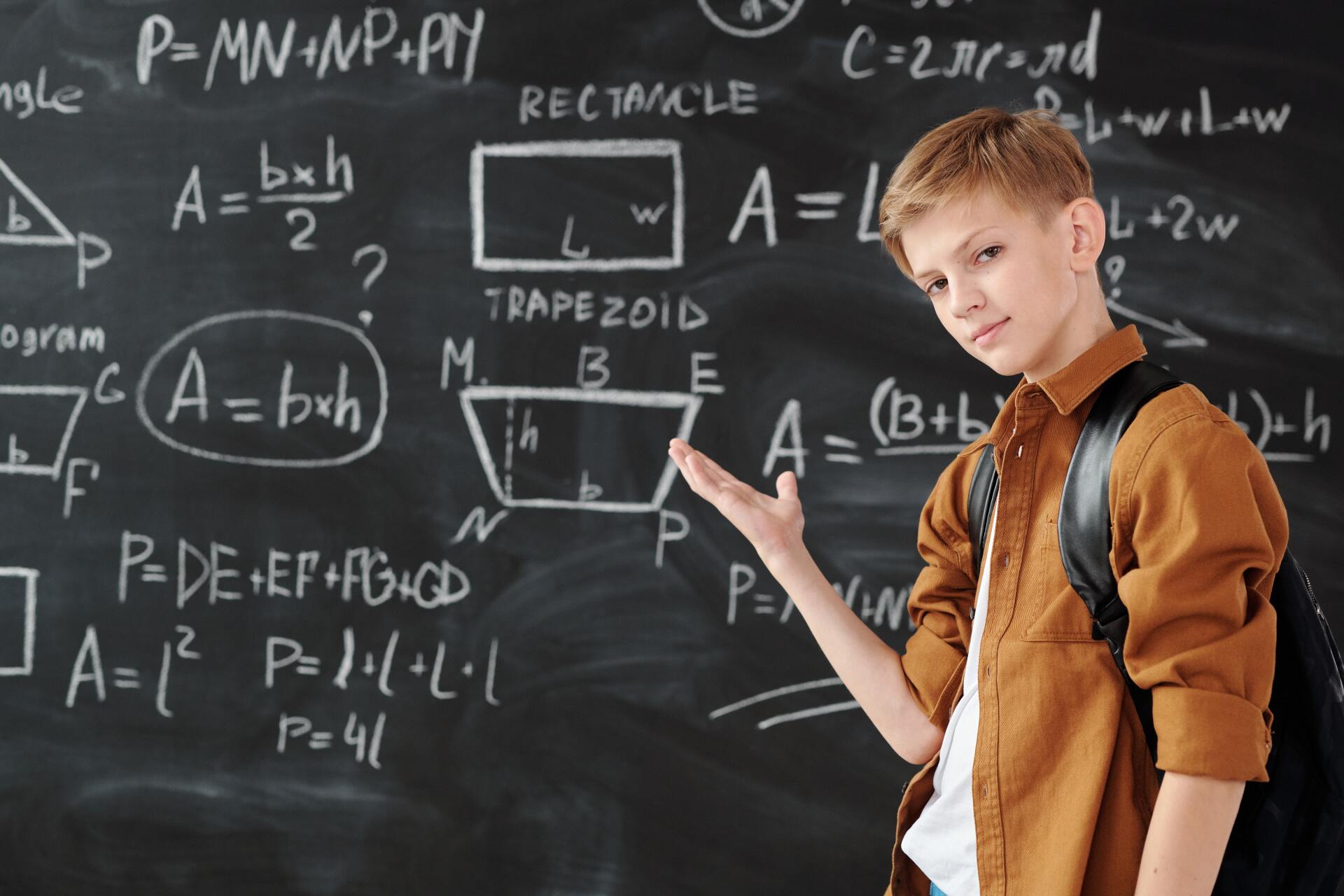 Young student pointing to math formulas on a chalkboard. He wears a brown shirt and a backpack, in a classroom setting.