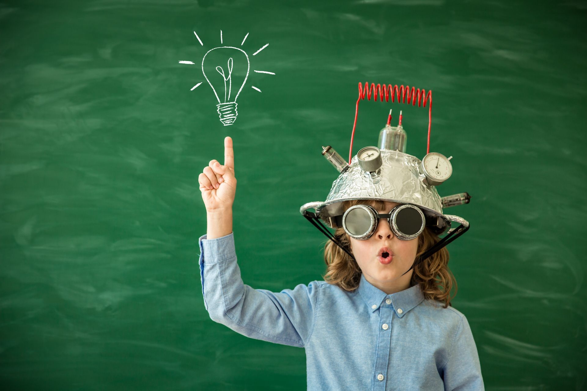 Child in a quirky inventor's hat pointing at a lightbulb drawing on a chalkboard, indicating an idea.