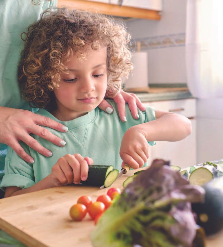 Child slicing zucchini with guidance from a person's hands in a kitchen, vegetables on a cutting board.