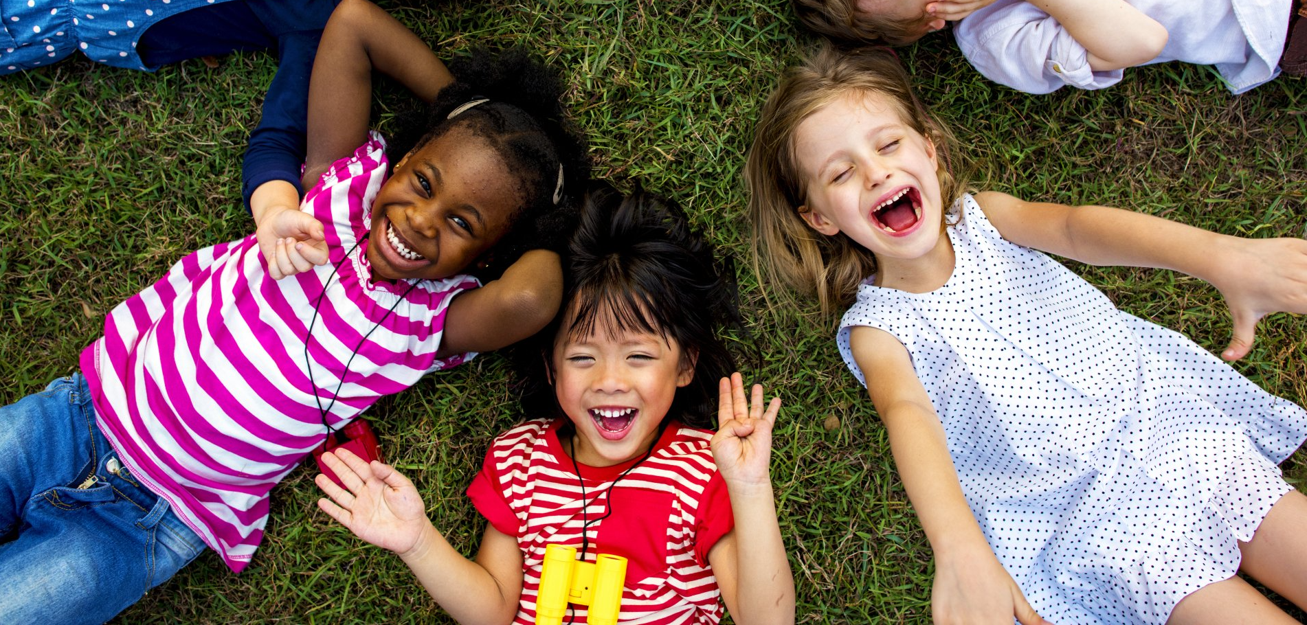 Four smiling children of diverse ethnicities lying on grass, laughing joyfully. Two children are wearing red striped shirts.