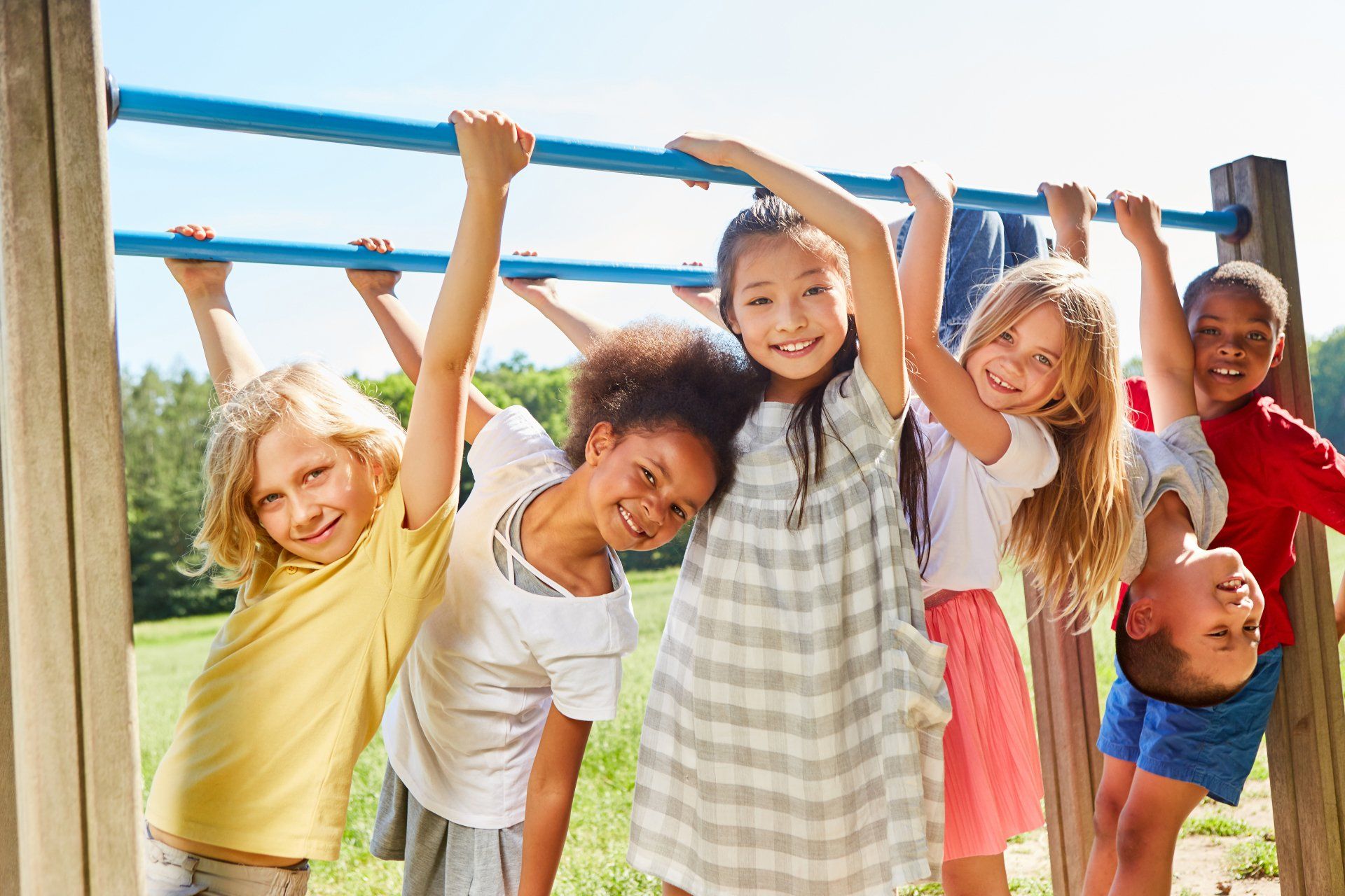 Group of smiling children hanging from blue monkey bars at a playground. Diverse group with different skin tones and expressions.