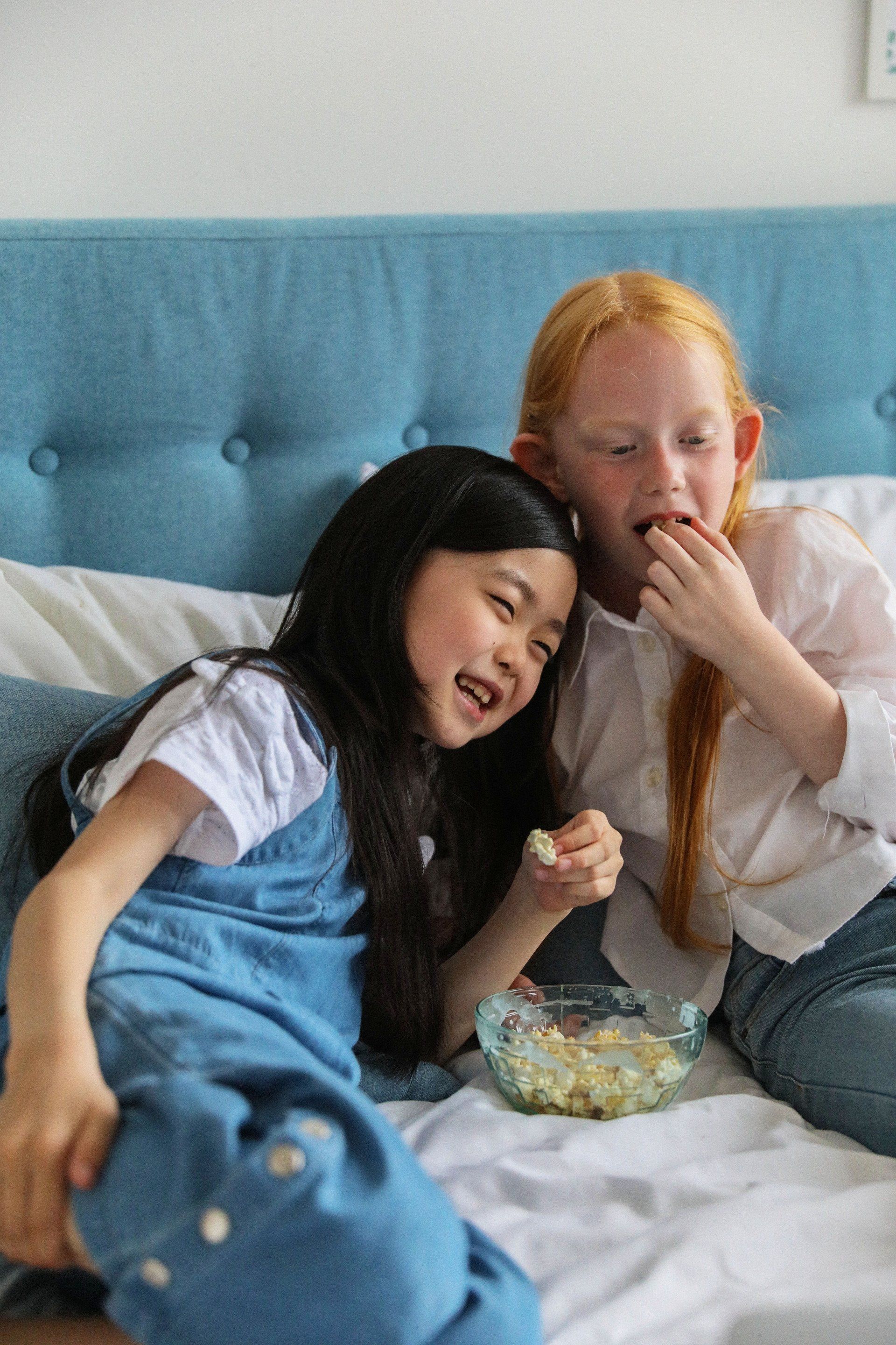 Two young girls, one with black hair and the other with red hair, eat popcorn on a bed with a blue headboard, smiling.