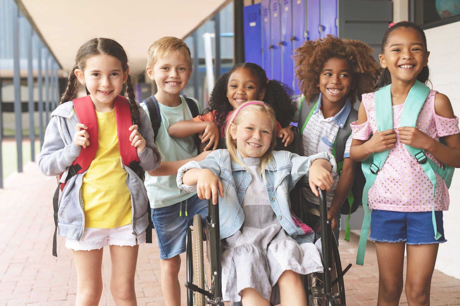 A group of smiling school children with backpacks, one using a wheelchair, pose in a school hallway.