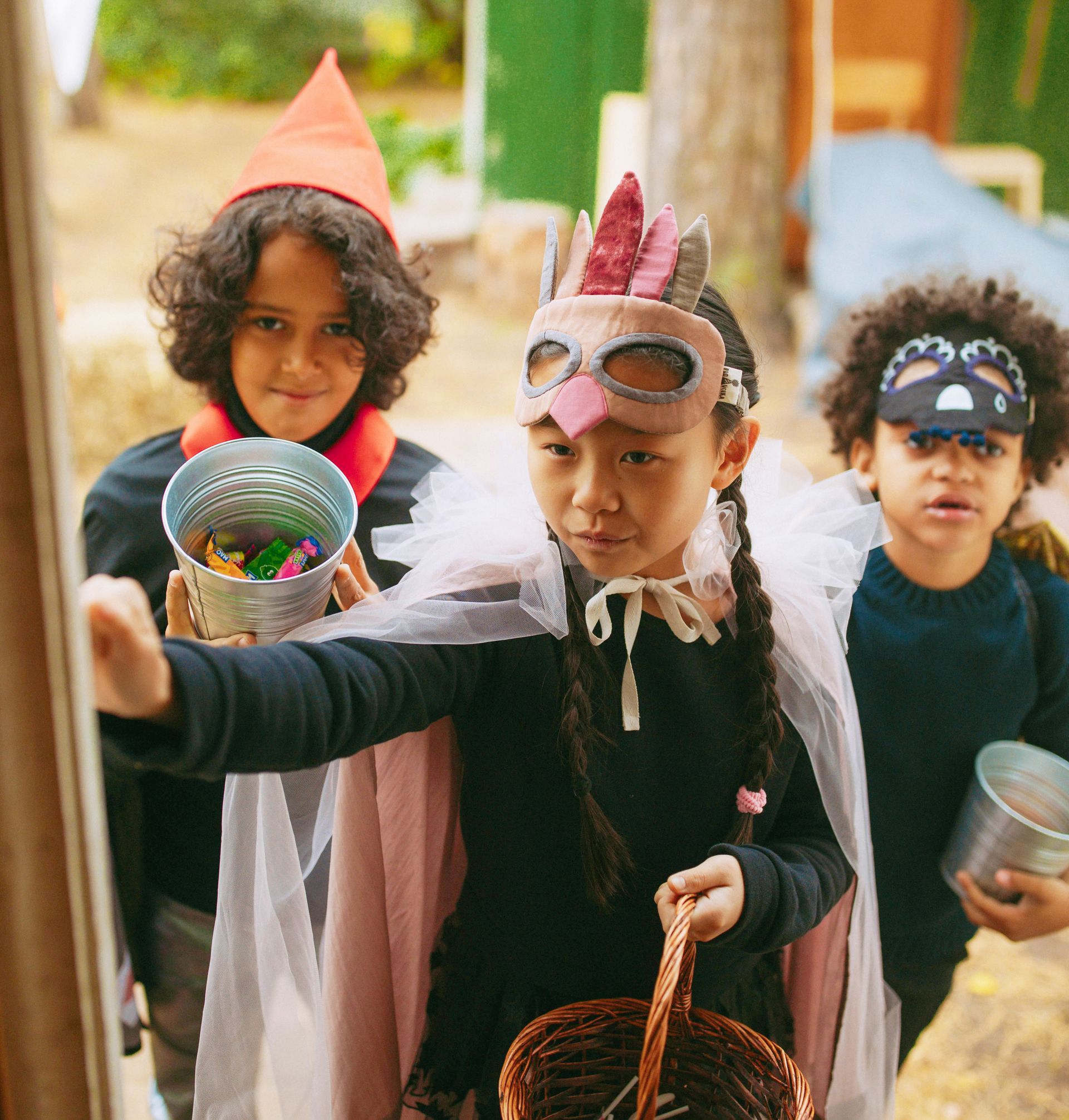 Three children in Halloween costumes trick-or-treating at a doorway. One holds a basket, one a pail of candy, and the other a container.
