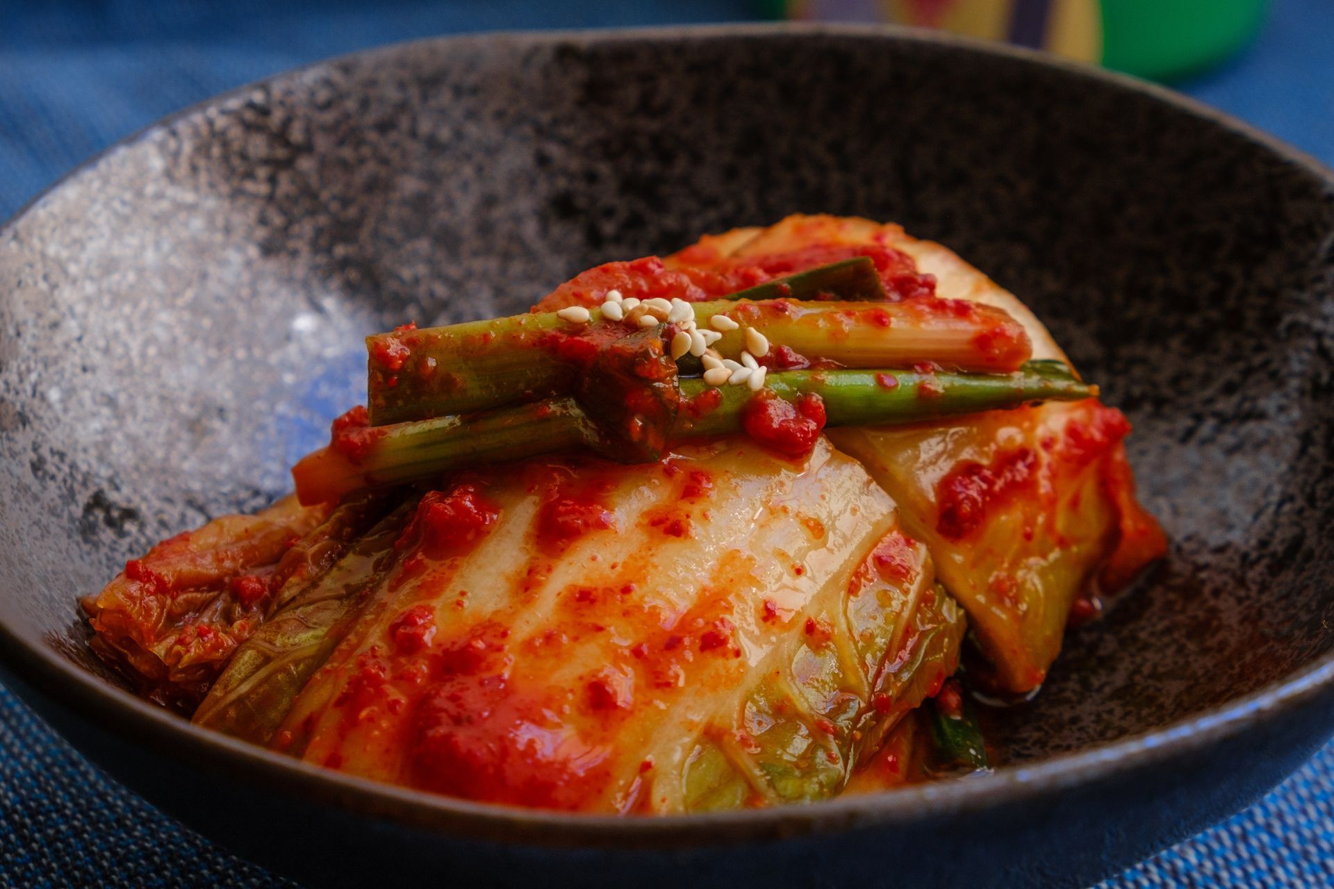 Close-up of kimchi in a dark bowl. The fermented cabbage is bright red with green onion and sesame seeds.