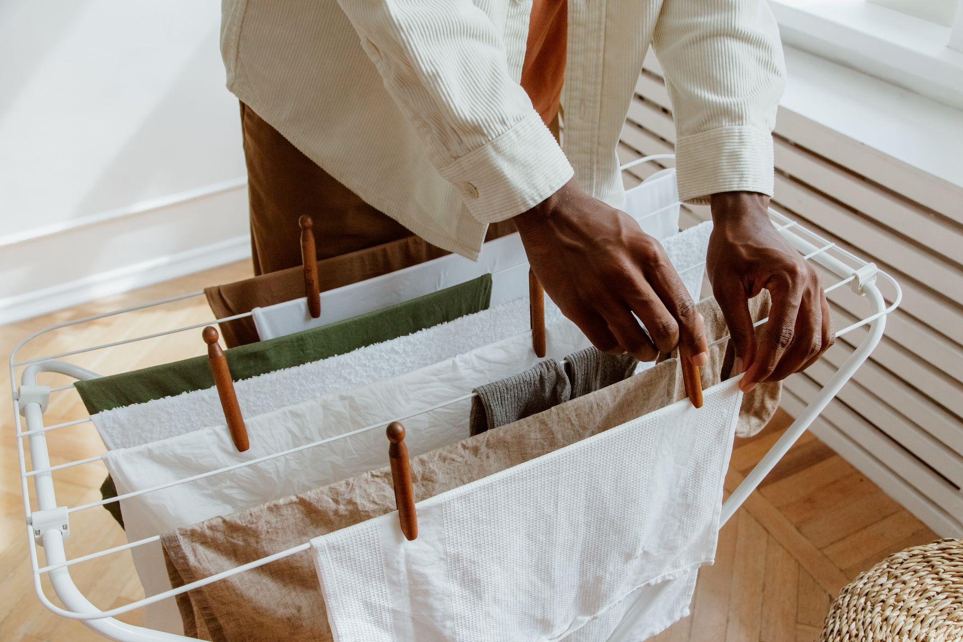 Person hanging laundry on a drying rack indoors; white, tan, and olive colored towels are clipped with wooden clothespins.