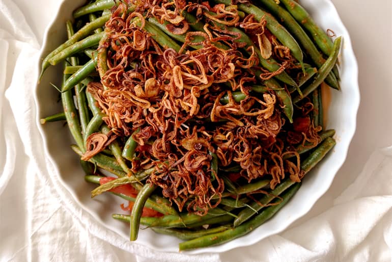 Green beans topped with crispy fried shallots in a white bowl on a white tablecloth.