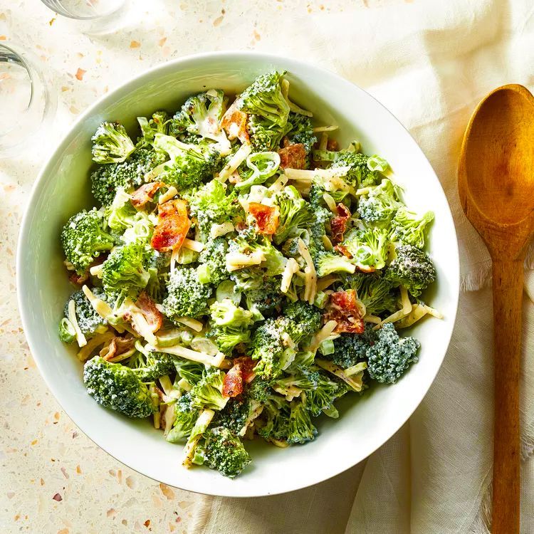 Broccoli salad in a white bowl with a wooden spoon. The salad contains broccoli florets, bacon bits, and a creamy dressing.