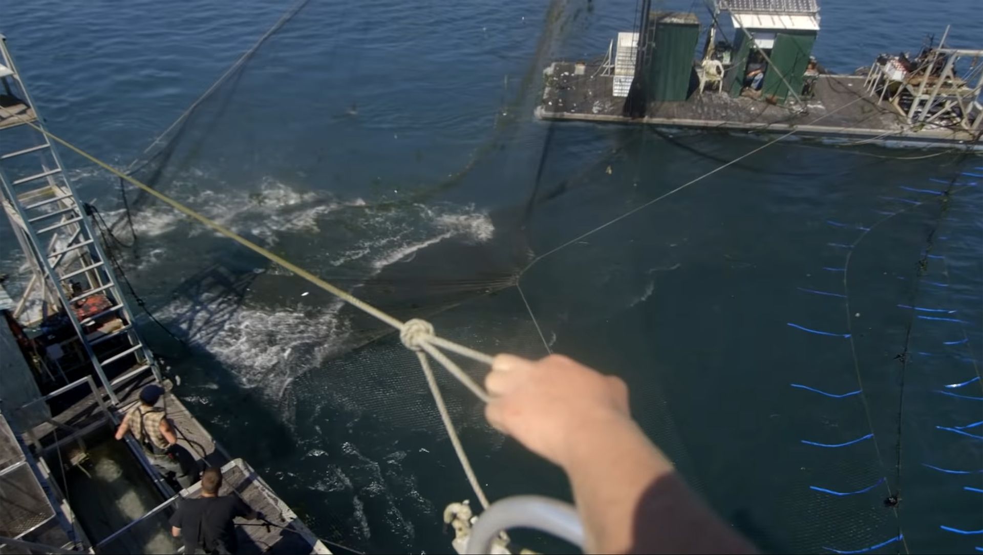 A fishing net being pulled up from the water by a boat, with another boat in the background.