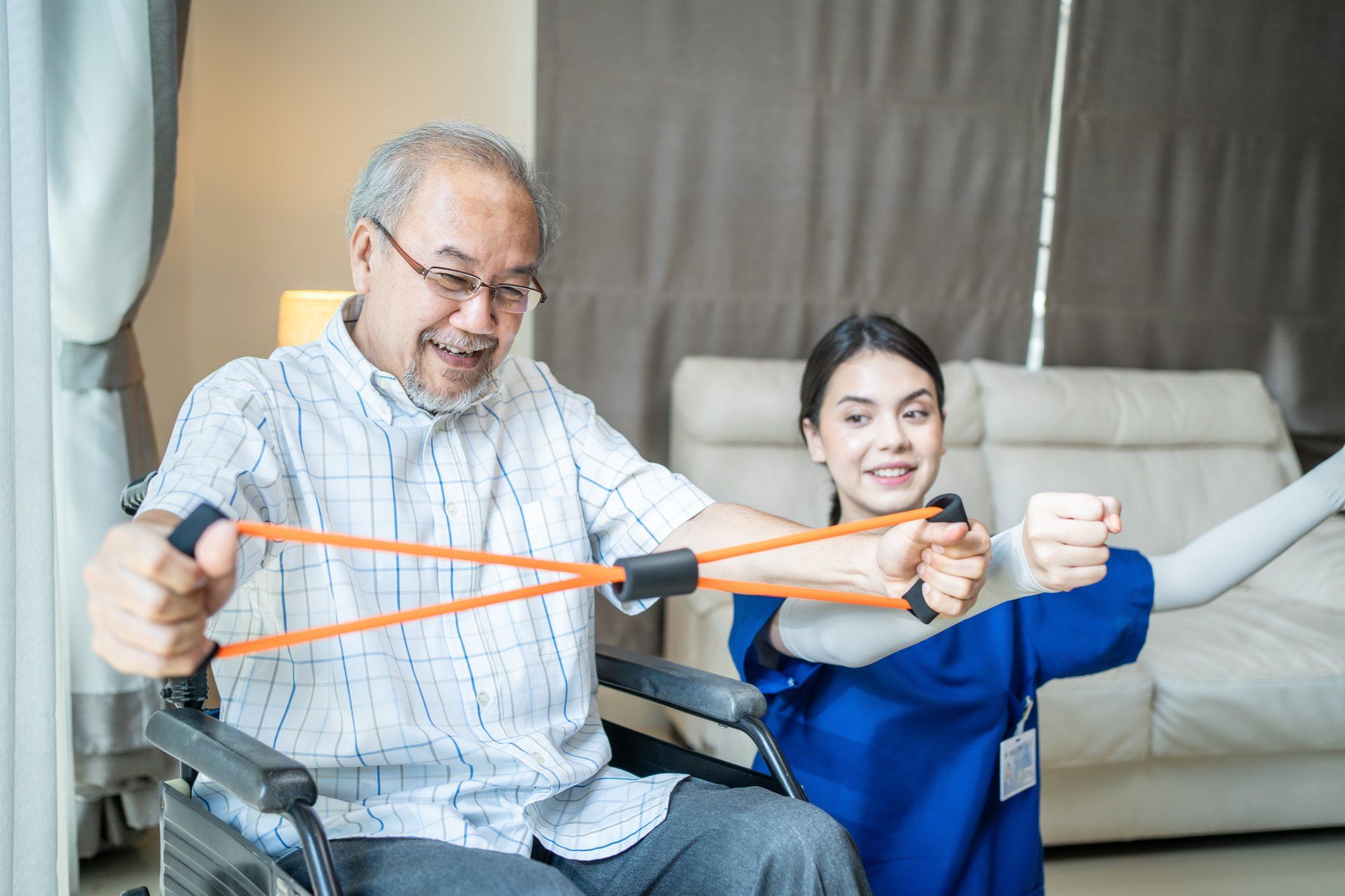 An elderly man in a wheelchair does resistance band exercises with the help of a smiling female caregiver in a room.