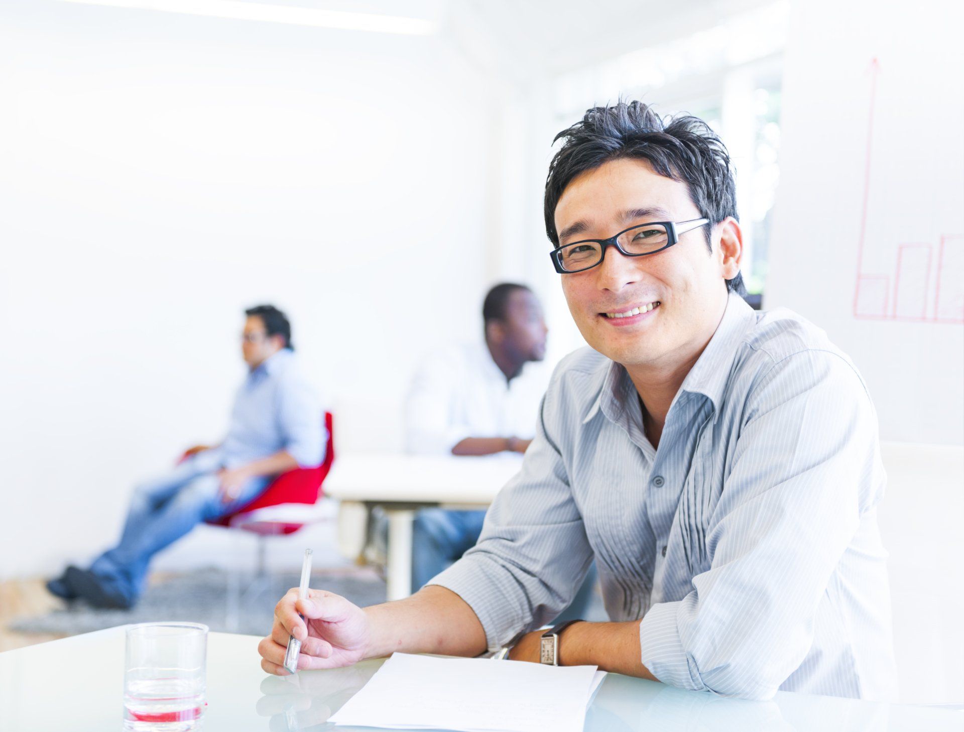 Smiling Asian man wearing glasses in a bright office, holding a pen and looking at the camera. Two colleagues in the background.