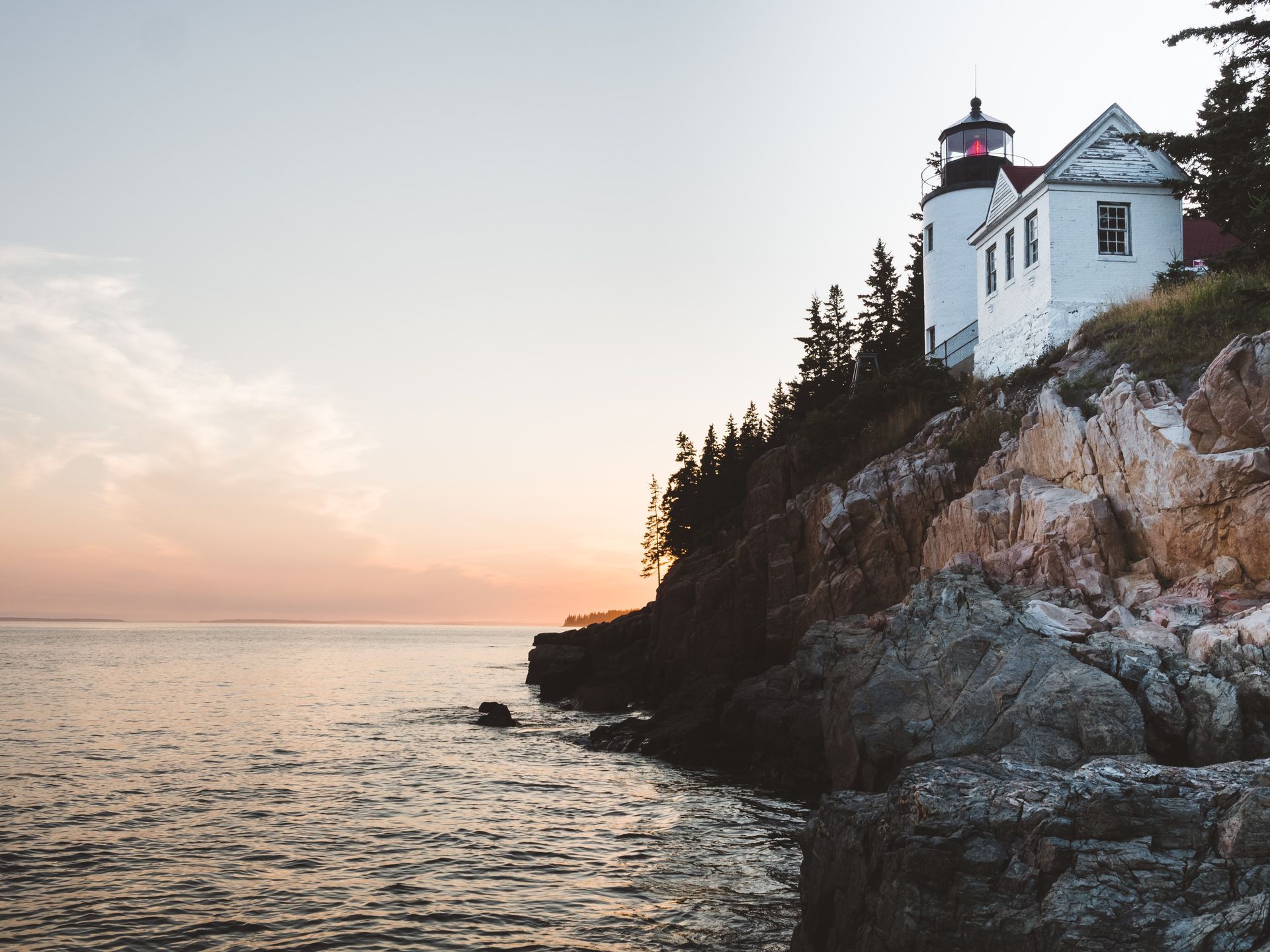 Lighthouse perched on rocky cliffside at sunset, with the ocean in the foreground and a dusky sky.