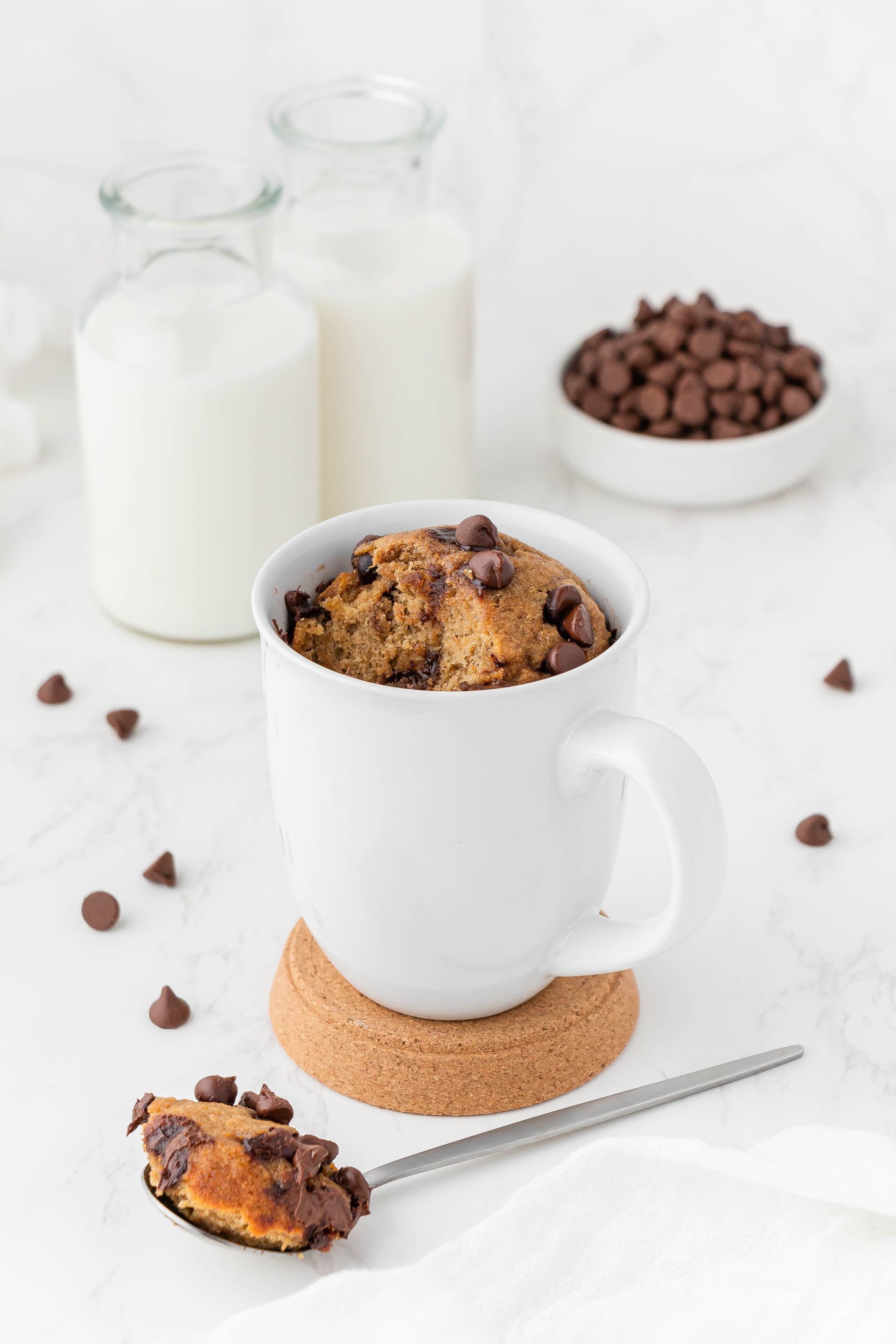 Chocolate chip mug cake in a white mug on a cork coaster, spoon in front with a bite, milk bottles and chocolate chips in the background.