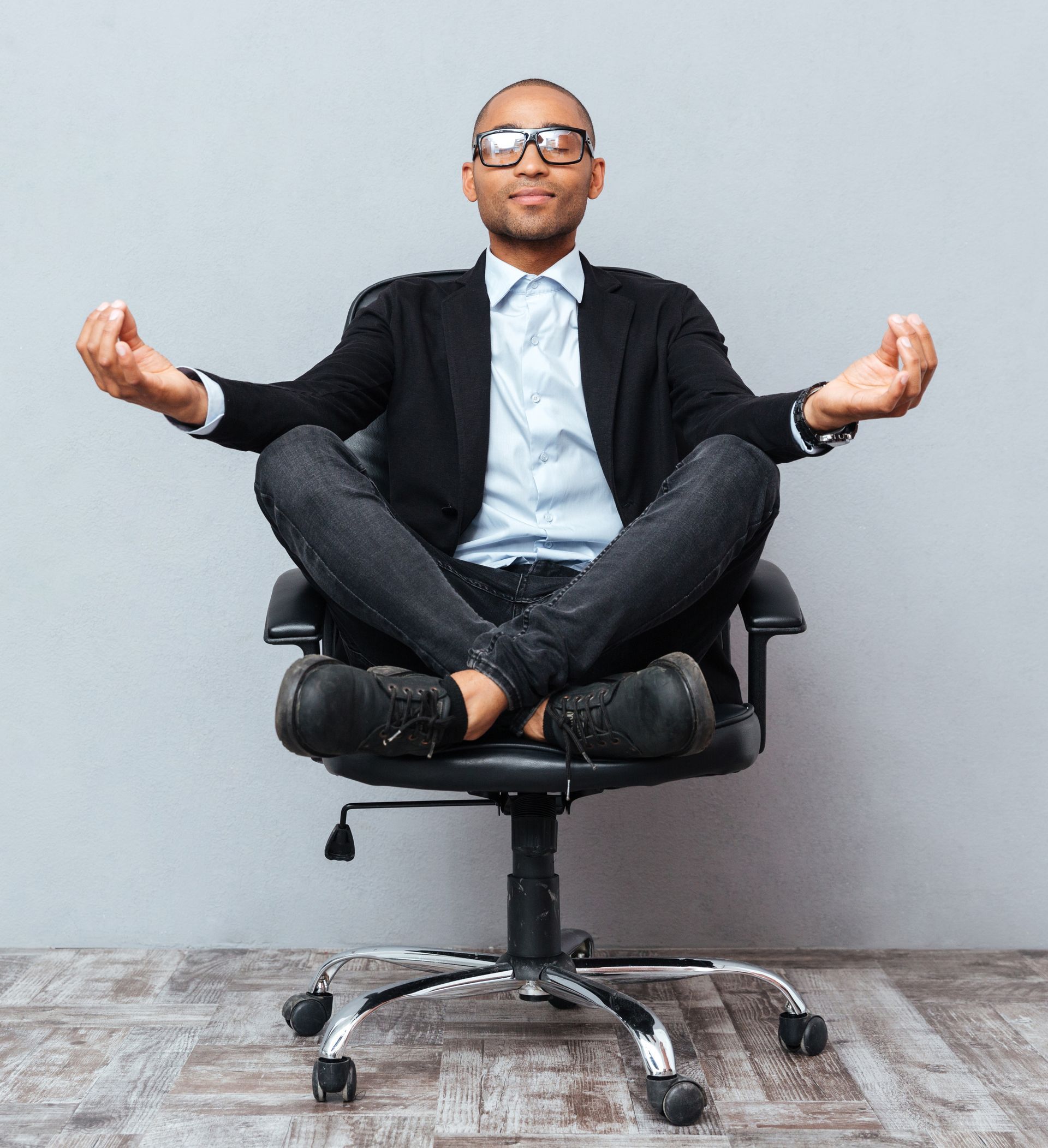 Black man in glasses meditating cross-legged on a chair; wearing a suit, gray backdrop.