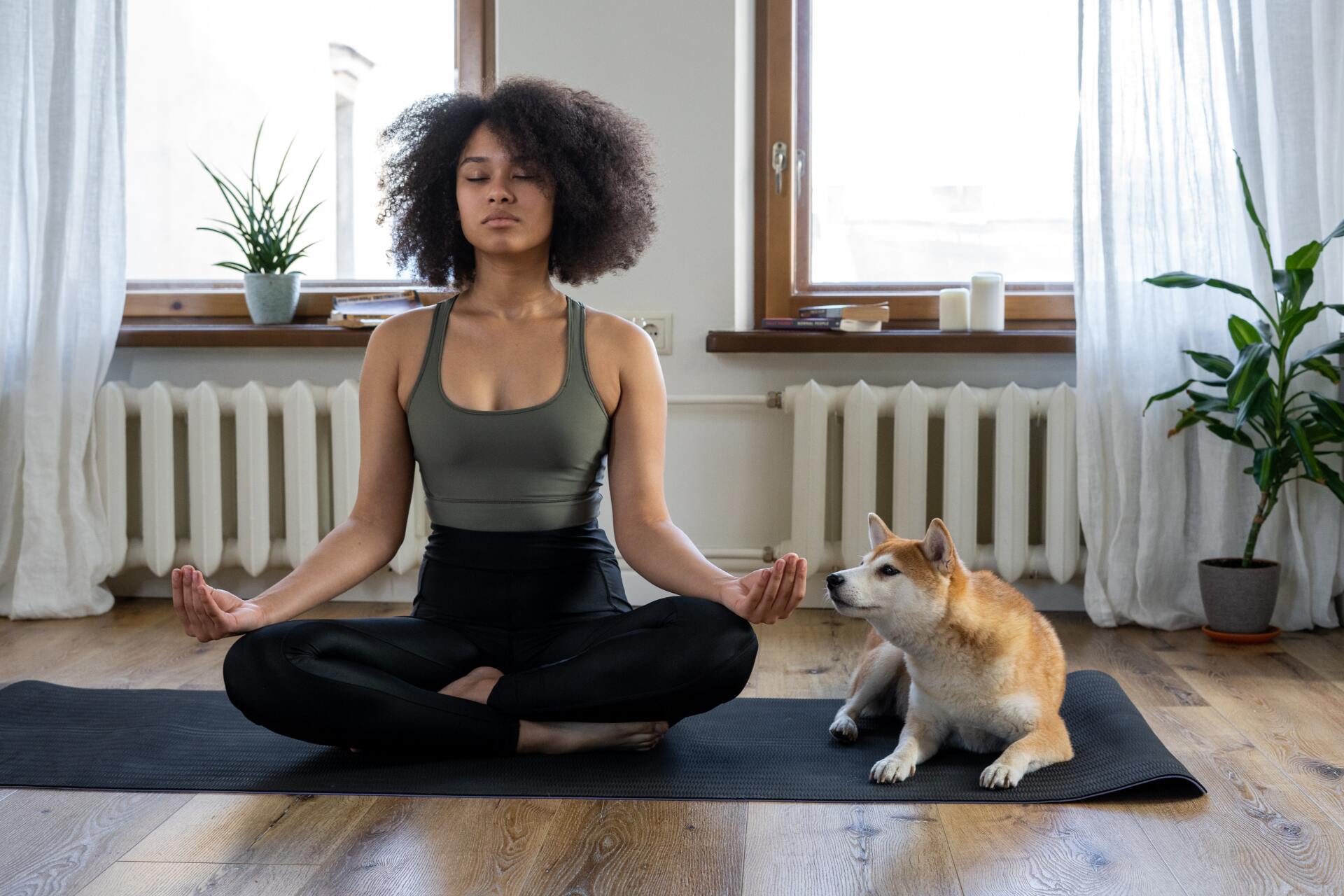 Woman with curly hair meditates on a mat indoors, eyes closed. A Shiba Inu dog sits nearby.