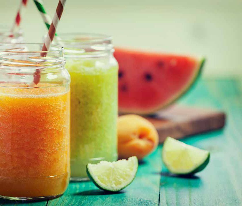 Colorful fruit smoothies in glass jars with straws, and slices of watermelon, lime, and apricot on a blue wooden surface.