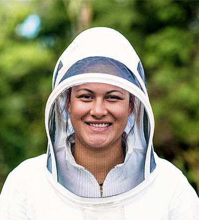 Smiling woman in a white beekeeping suit with hood and mesh face covering, against a blurred green background.