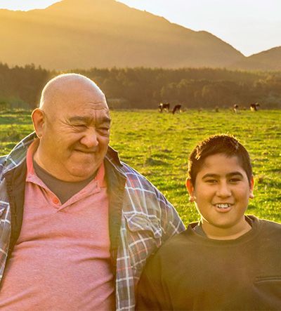 A smiling older man and a young boy stand together in a sunny field with mountains in the background; cows graze in the distance.