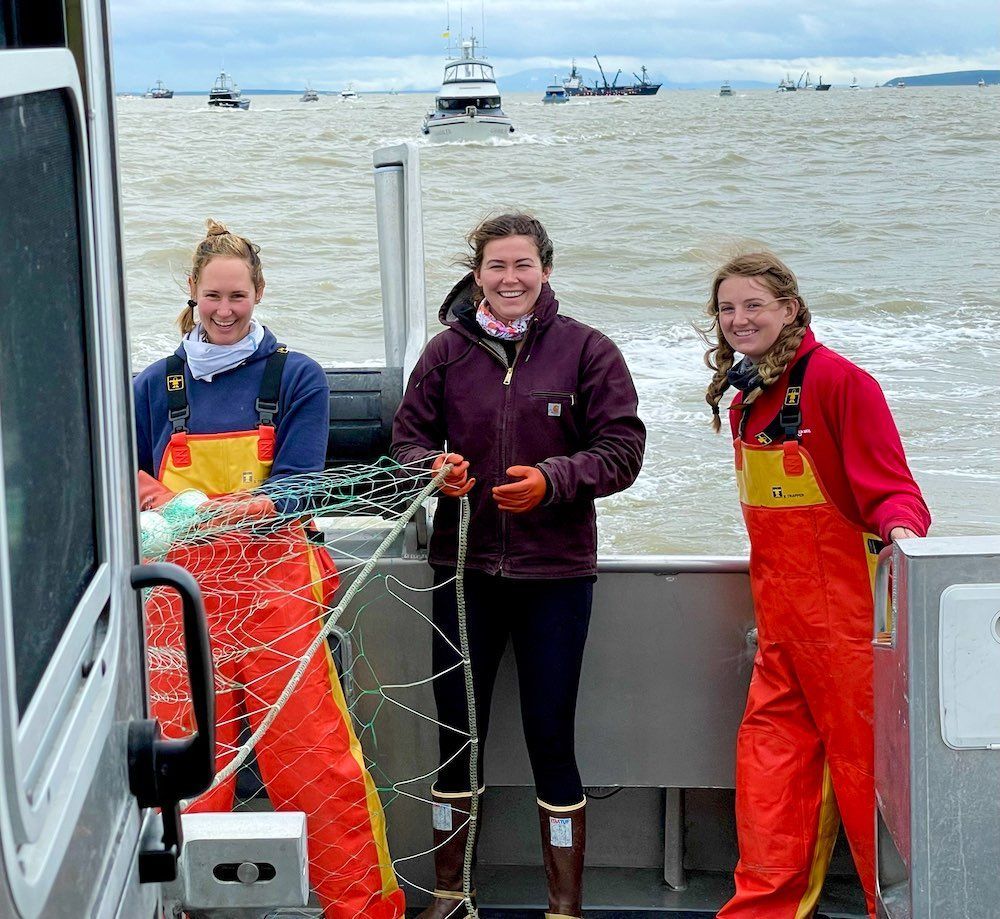 Three women on a boat, smiling. Two are holding crab traps, wearing orange overalls. Ships and cloudy sky in the background.