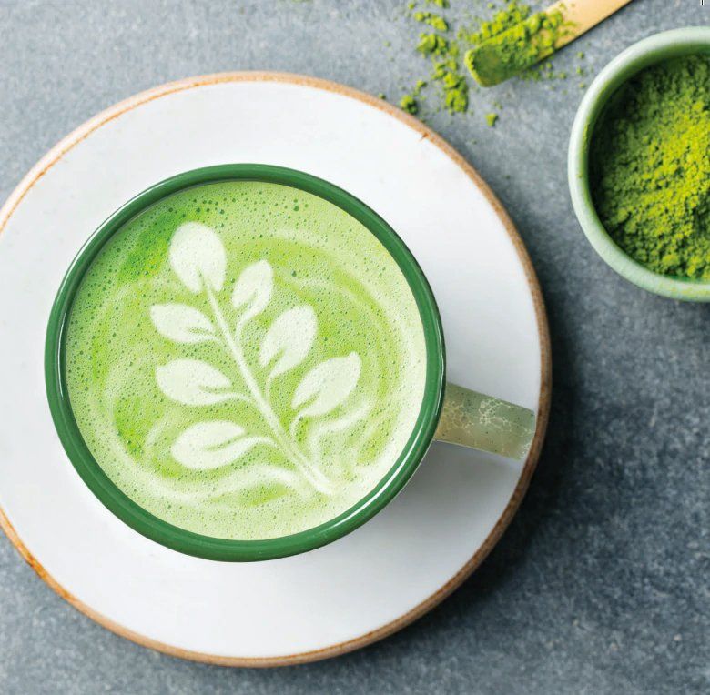 Matcha latte in a green-rimmed mug with leaf art. Next to it, a bowl and scoop of matcha powder.