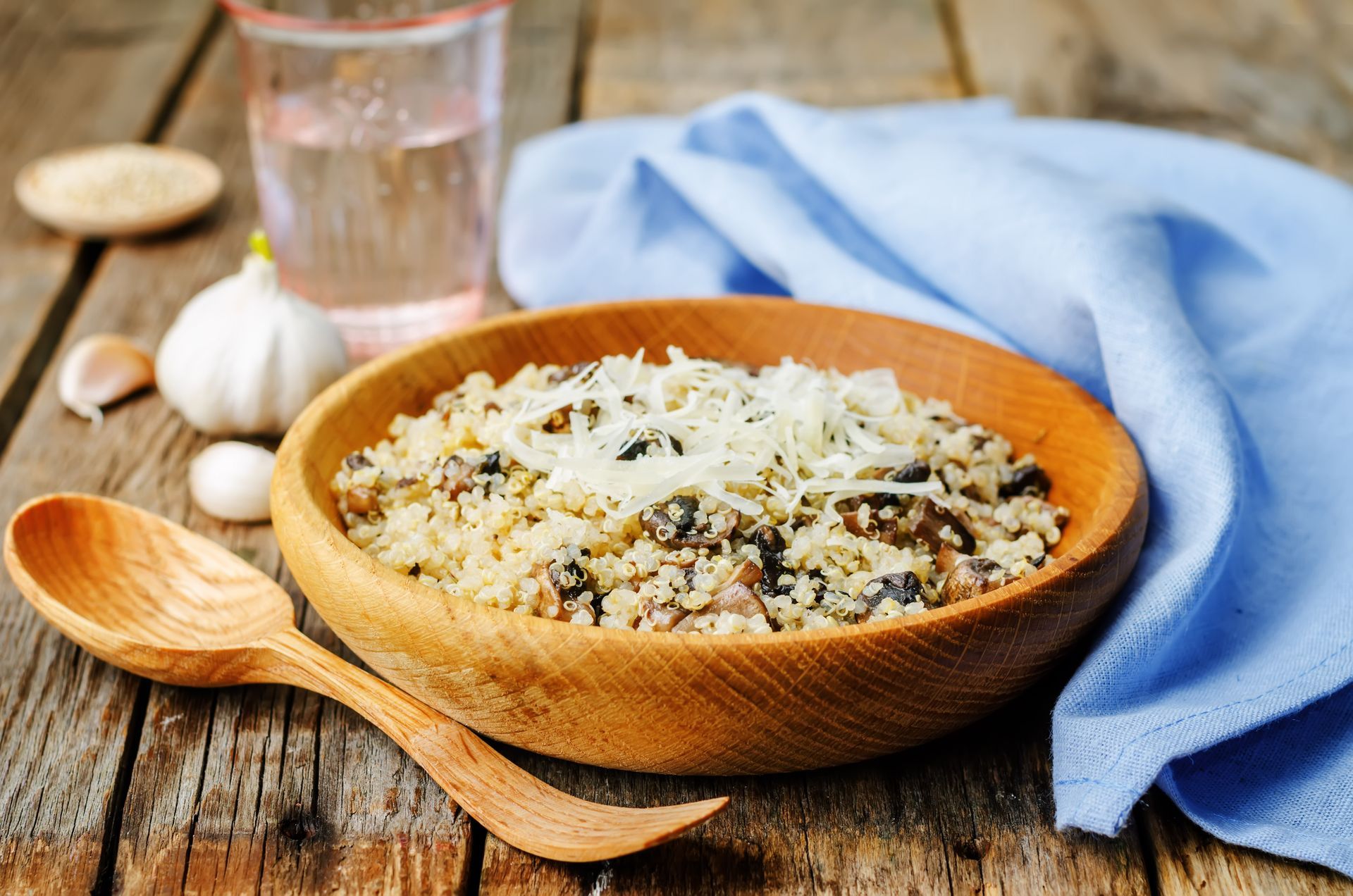 A wooden bowl of mushroom risotto topped with grated cheese, alongside a wooden spoon, garlic, and a glass of water.