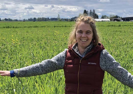 Woman with arms outstretched in a green field smiles. She wears a maroon vest and gray sweater under a cloudy sky.