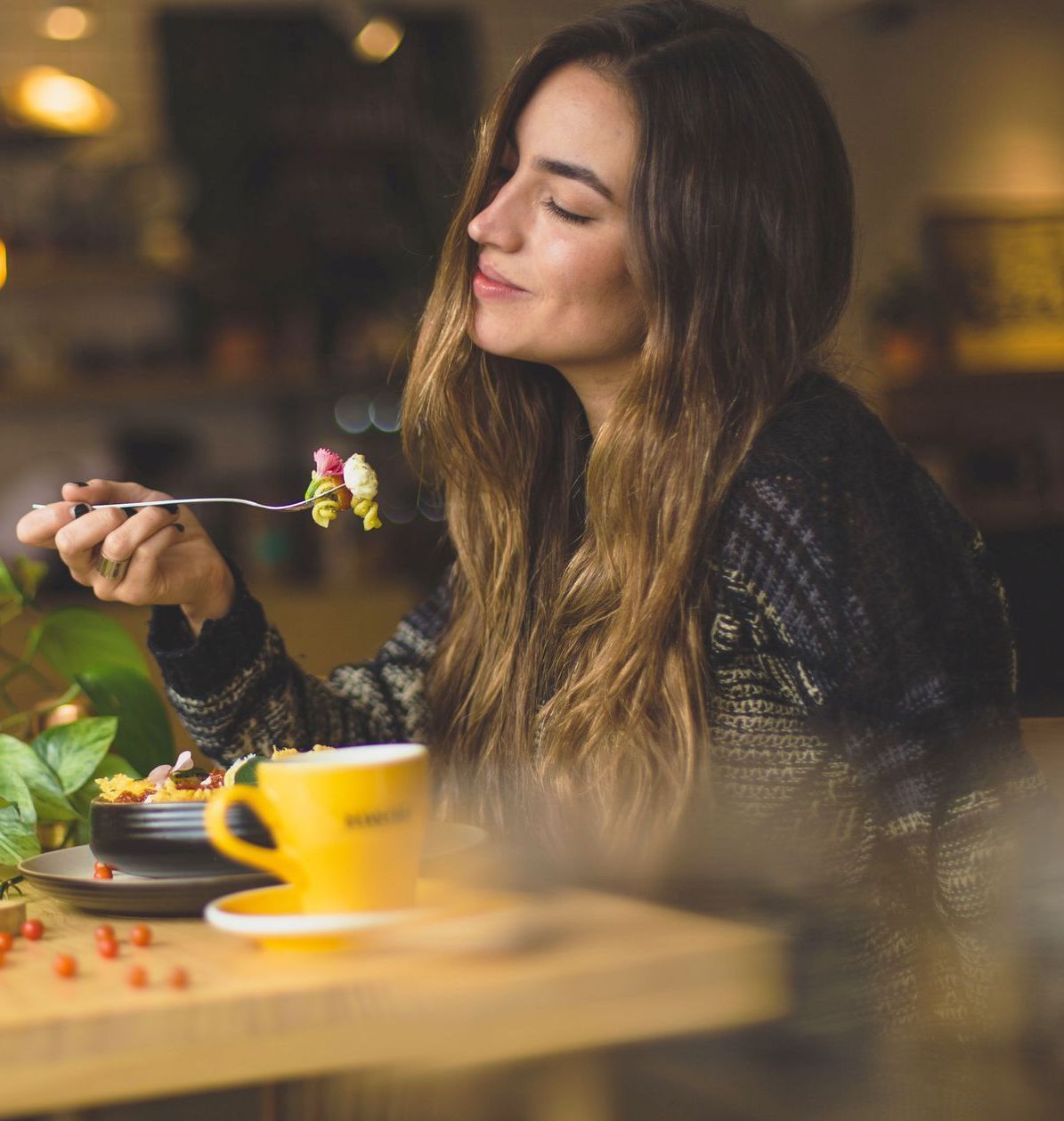 Woman enjoys food, smelling it with eyes closed in a cafe setting. She has long brown hair and is wearing a dark sweater, yellow cup on the table.