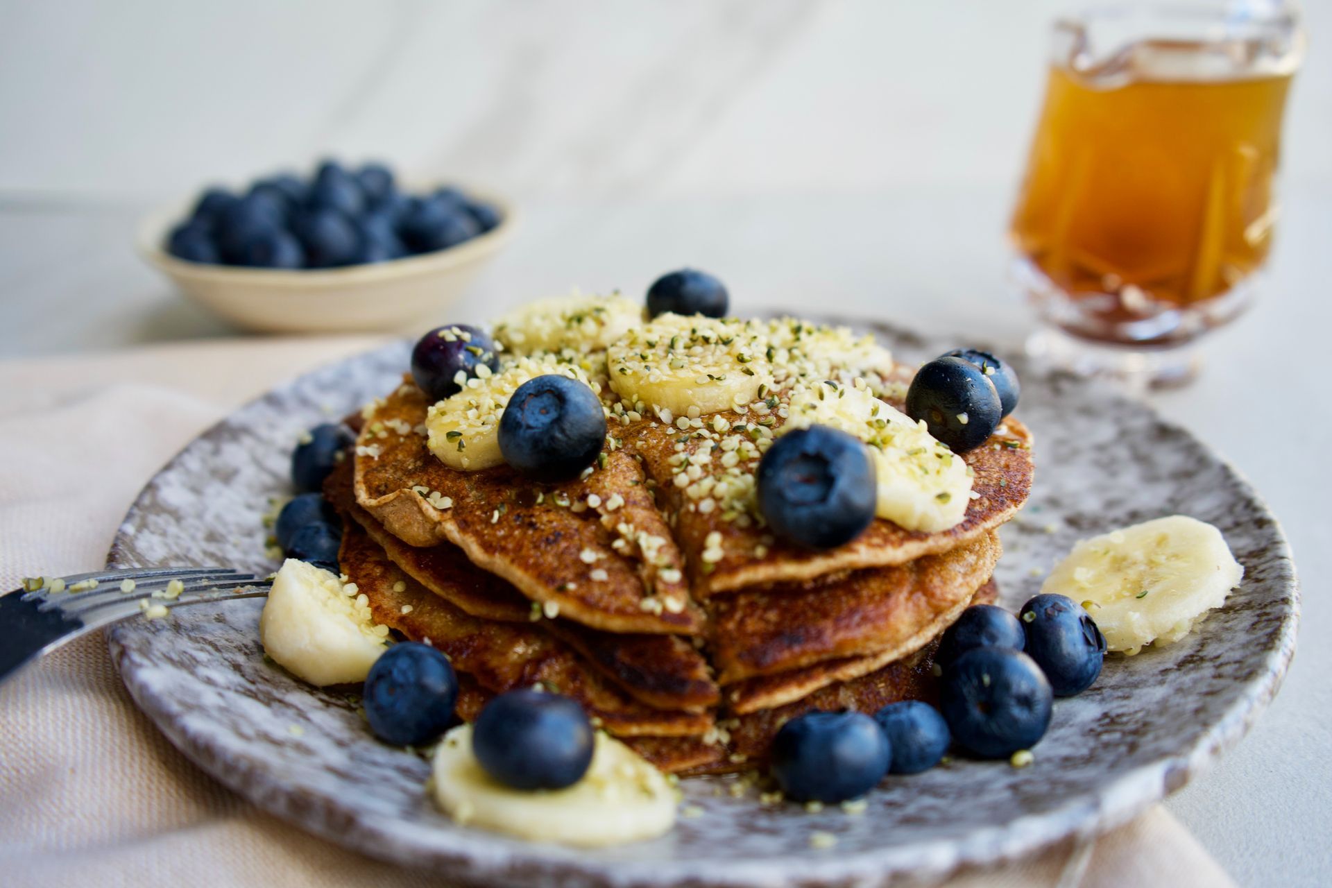 Stack of pancakes topped with blueberries, sliced bananas, and sprinkled with hemp seeds, served on a speckled plate with a side of maple syrup and a bowl of blueberries.