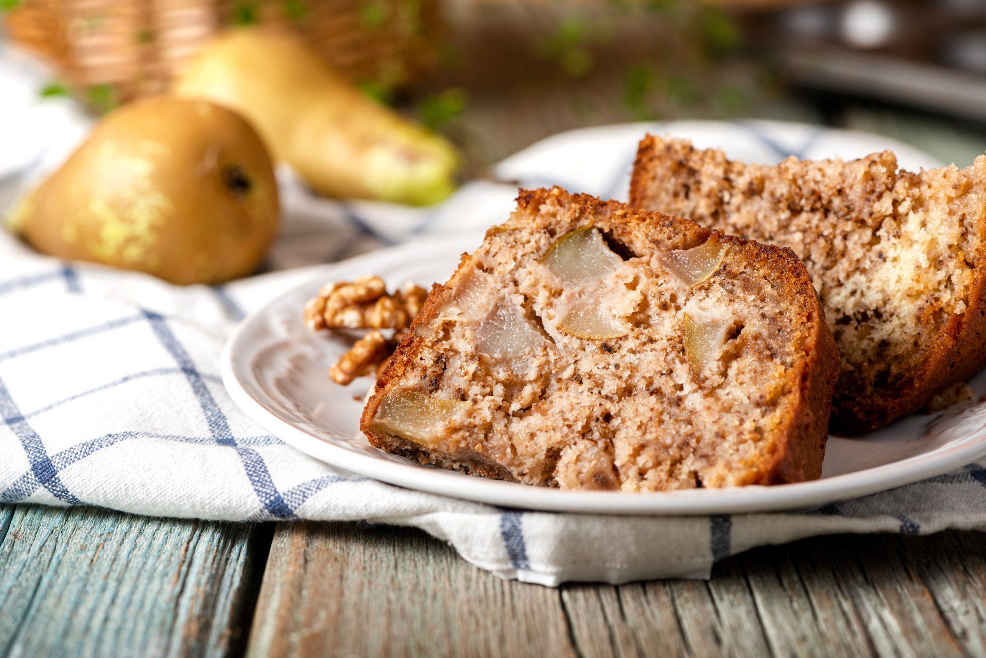 Pear cake slices on a white plate, with a pear and walnuts on a checkered cloth, on a wooden table.