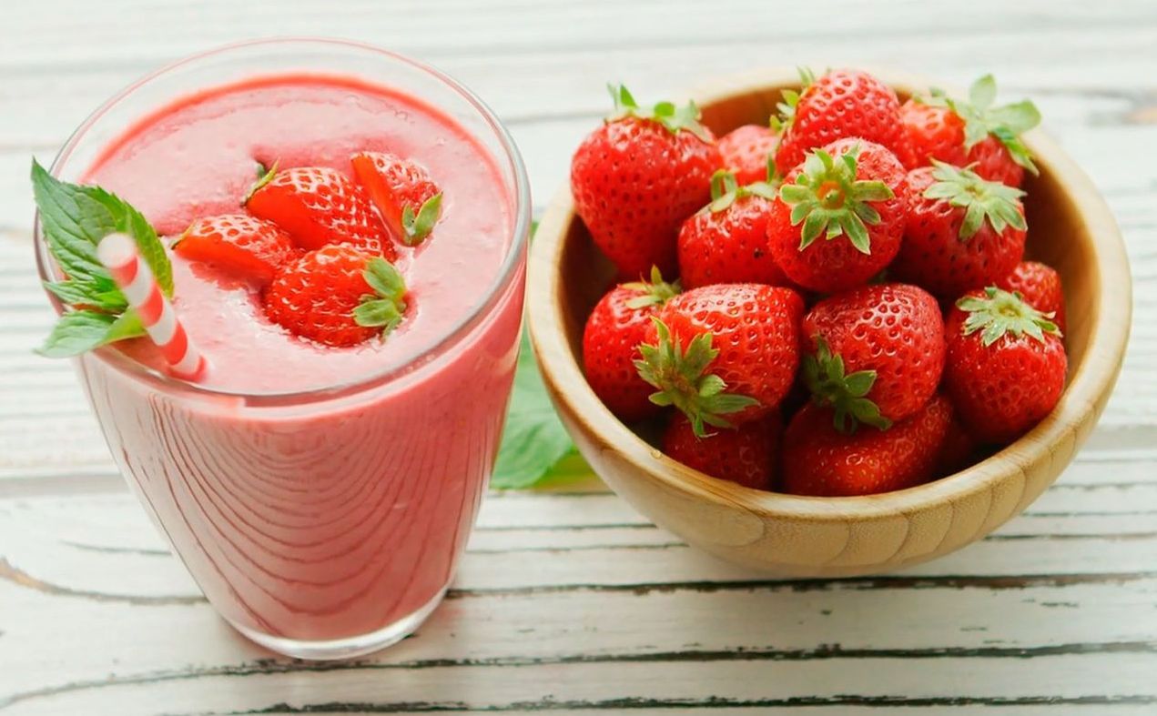 Strawberry smoothie in a glass, topped with fresh strawberries, beside a wooden bowl filled with strawberries on a white-washed wooden surface.
