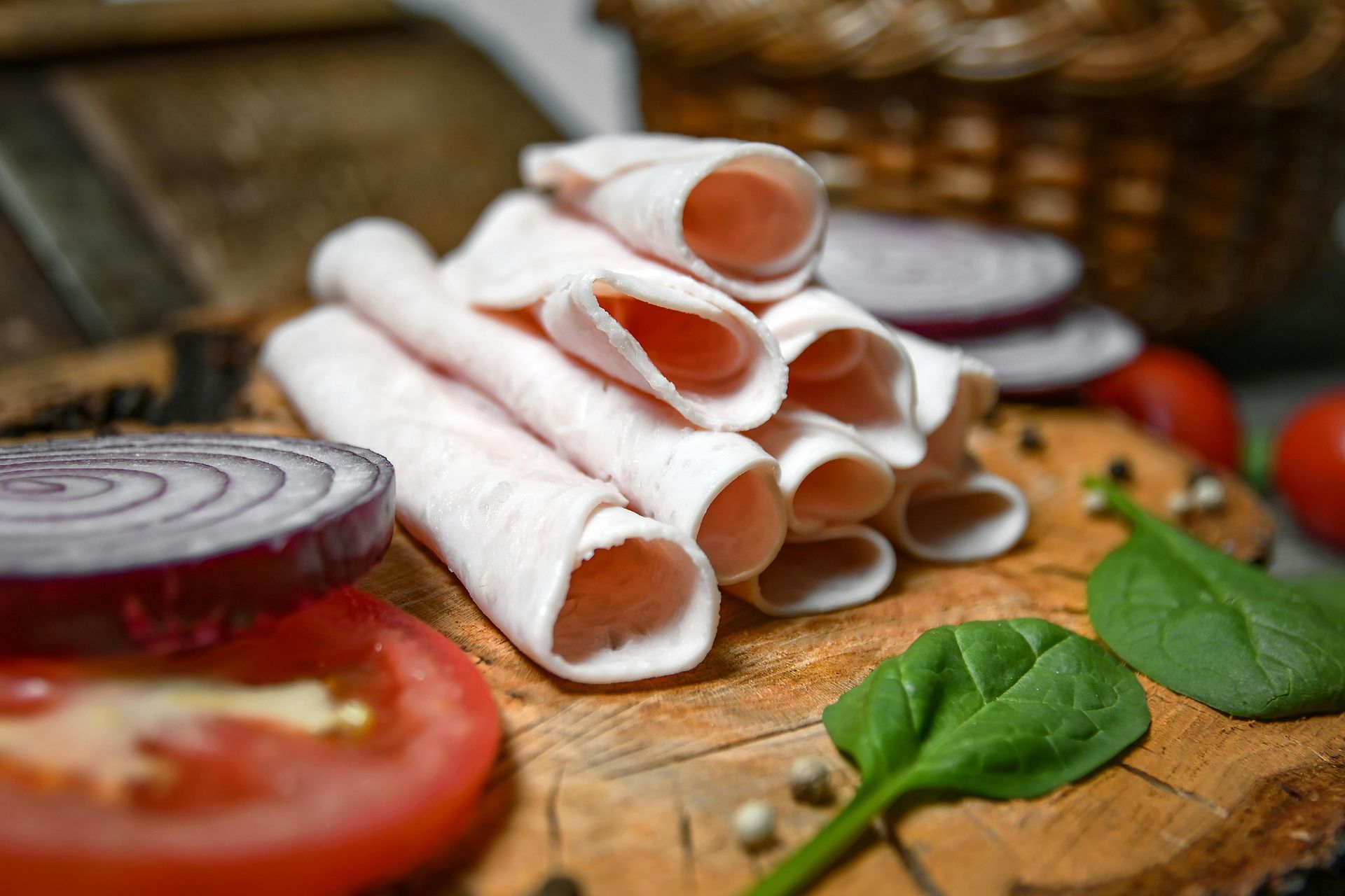 Sliced deli meat rolls stacked on wood, with tomato, onion, and spinach.