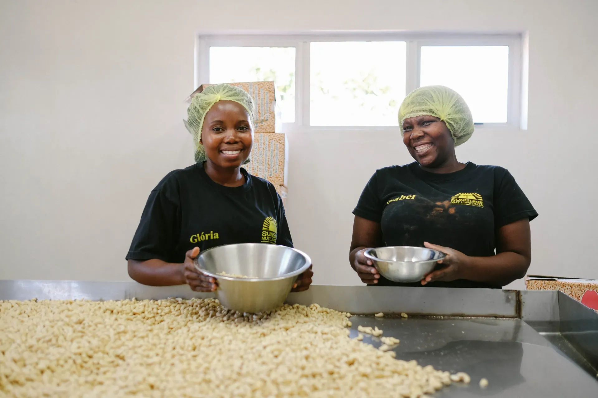 Two smiling Black women in hairnets sort coffee beans at a processing table. They hold bowls, one looking at the camera, the other smiling sideways.