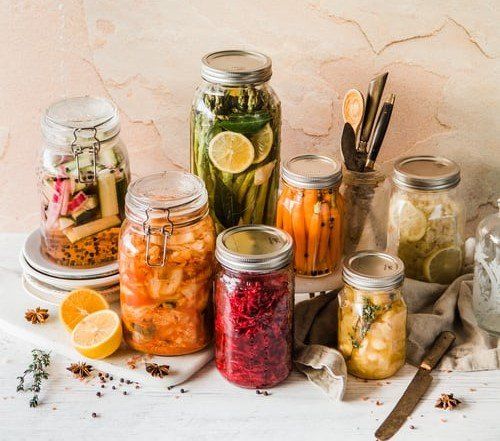 Jars of pickled vegetables in various colors like red, orange, and green sit on a white surface, with scattered spices and lemons.