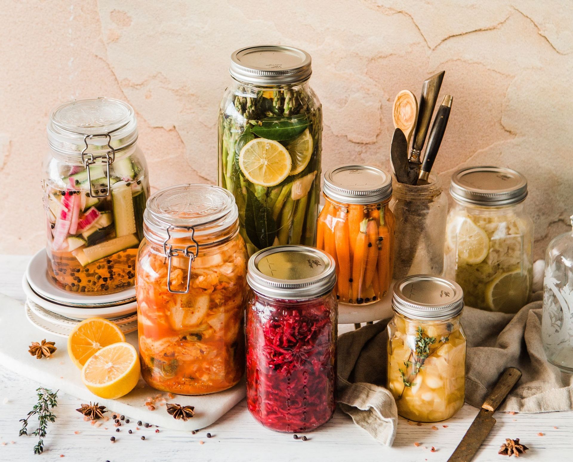 Various jars of pickled vegetables, including carrots, beets, asparagus, and lemons, on a white surface with a neutral background.
