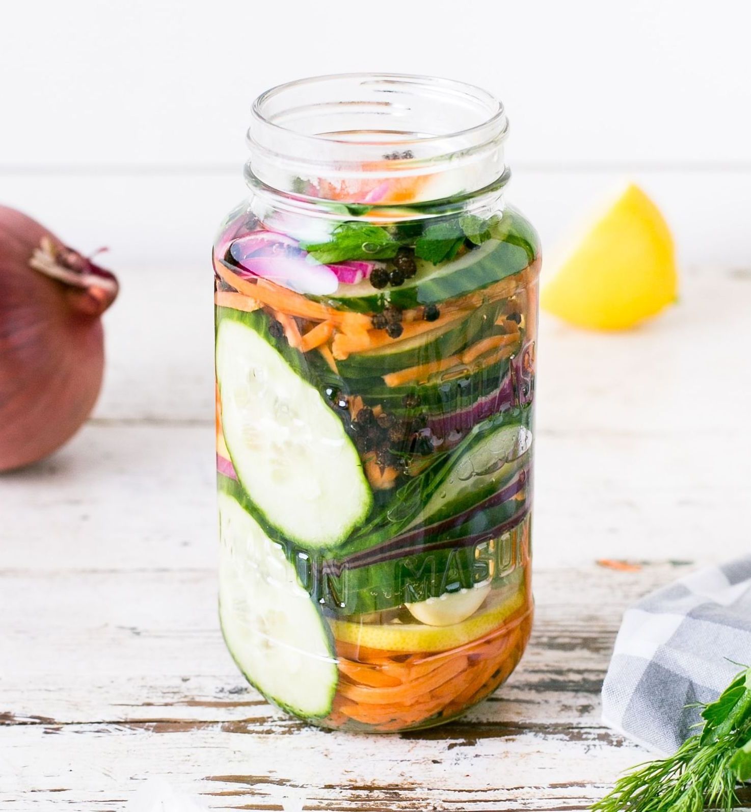 Jar of pickled vegetables with cucumbers, carrots, red onion, and herbs, with a red onion half and lemon wedge nearby.