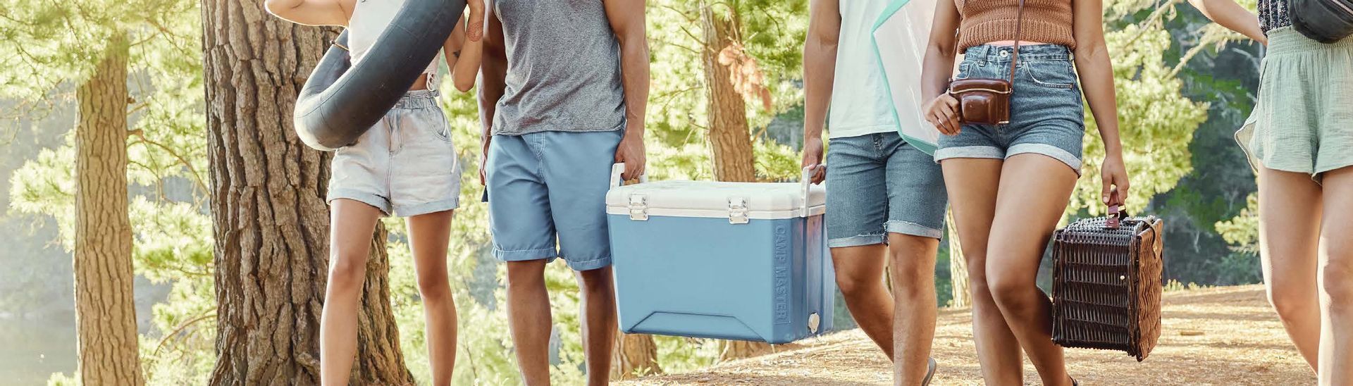 A group of friends walks along a tree-lined path carrying picnic supplies: cooler, bags, and a flotation device.